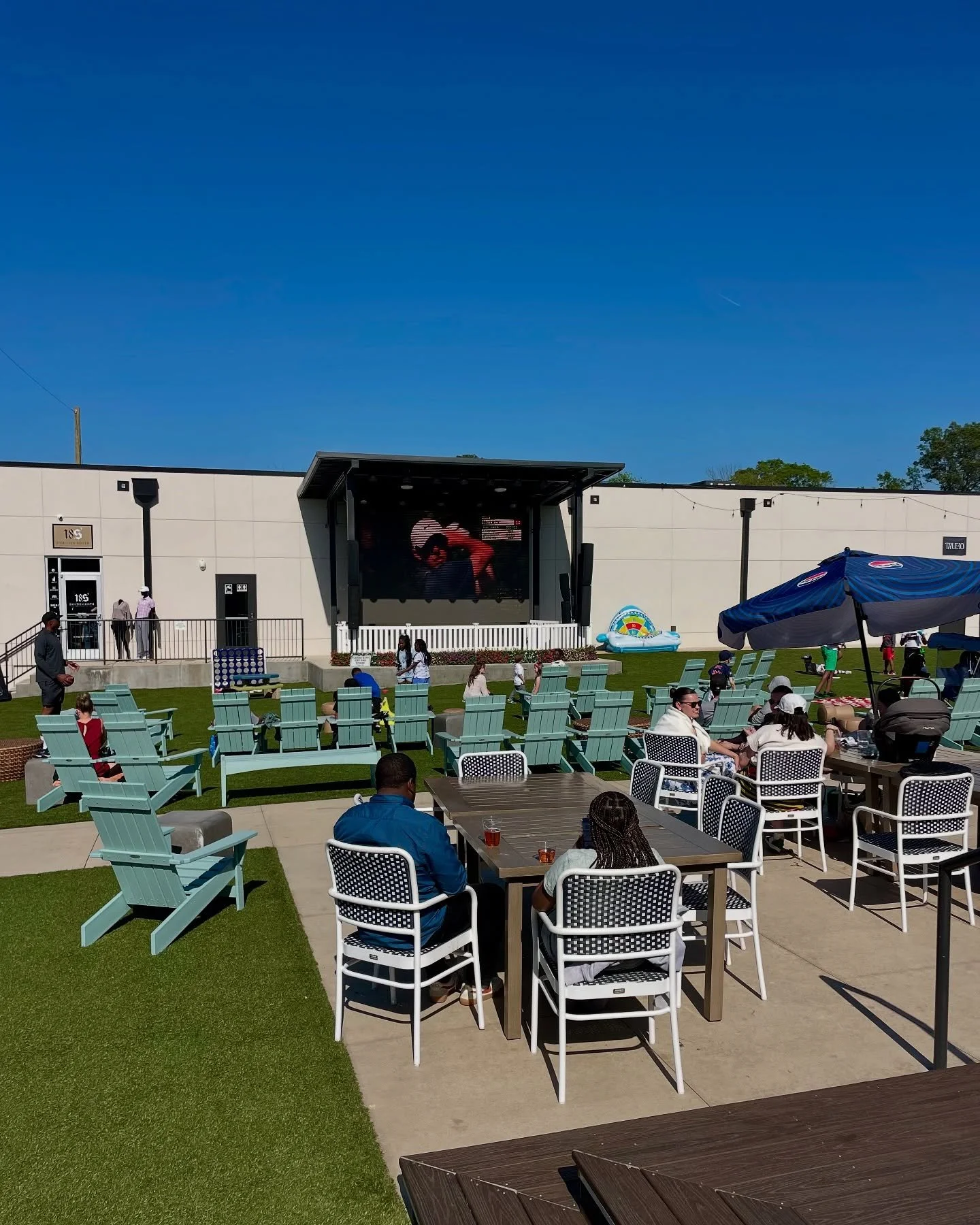 Outdoor patio area with tables, chairs, umbrellas, and a lawn where people are sitting and standing. A large screen on the building behind shows a person in a red outfit. There is a structure with a stage or screen on the building, and a small paddle boat float in the background. The sky is clear and blue.