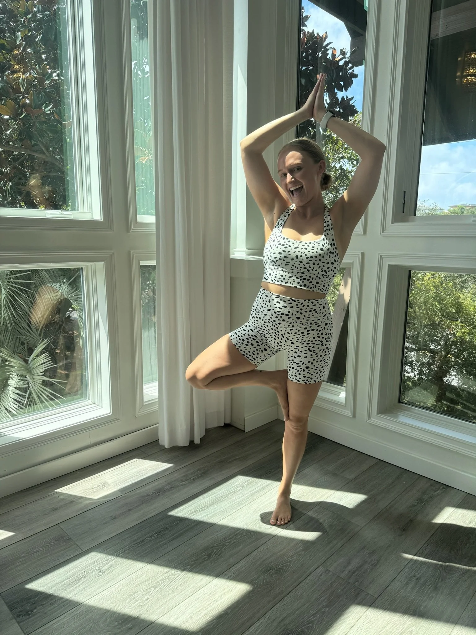 A woman performing a yoga pose in a sunlit room with large windows, wearing a black and white polka dot sports bra and matching shorts, standing on one leg with the other bent and foot resting on her thigh, smiling and raising her hands above her head with palms together.