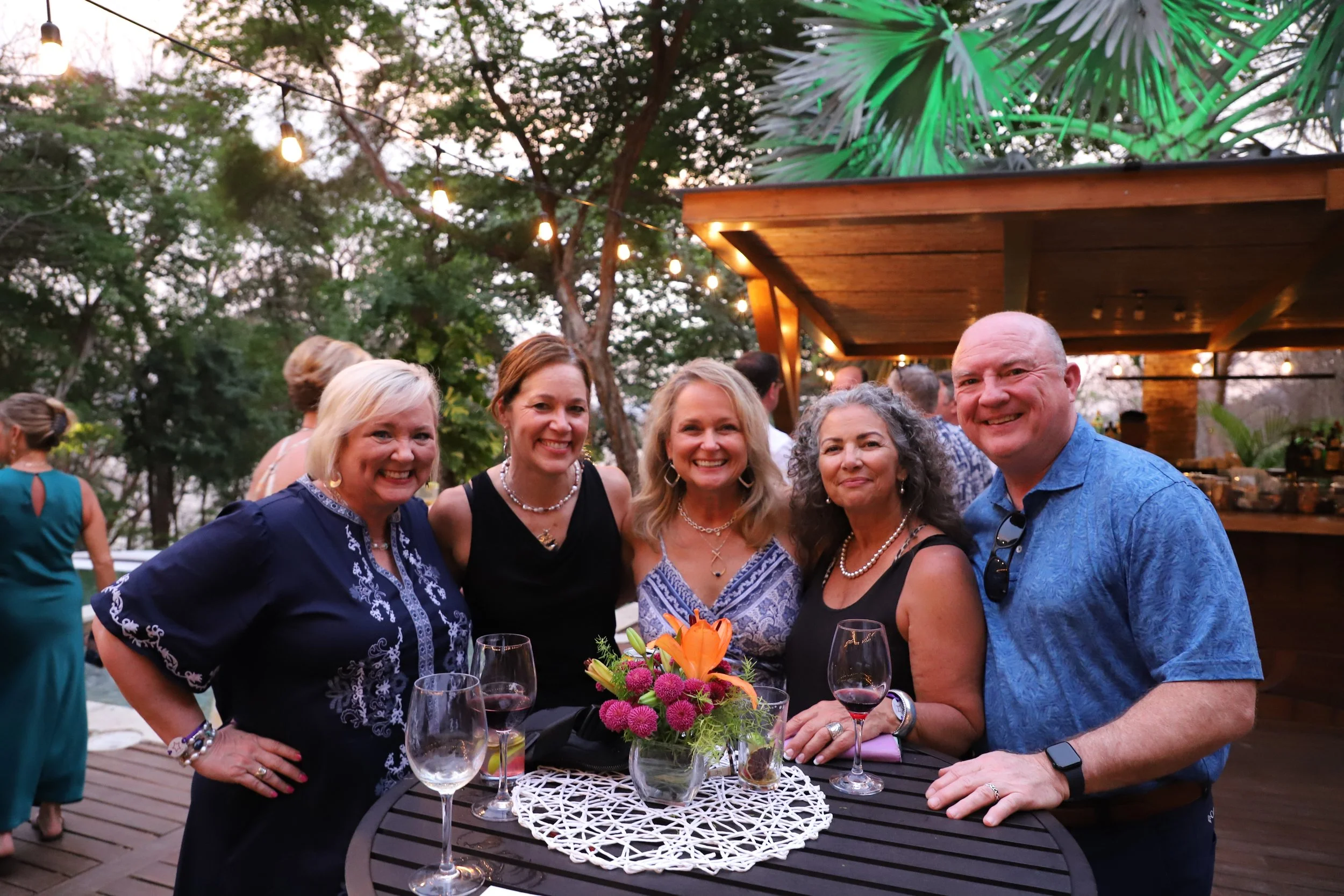 A group of five smiling adults, three women and two men, standing around a table at an outdoor evening gathering with string lights and lush greenery in the background. They have glasses of wine and a floral centerpiece on the table.
