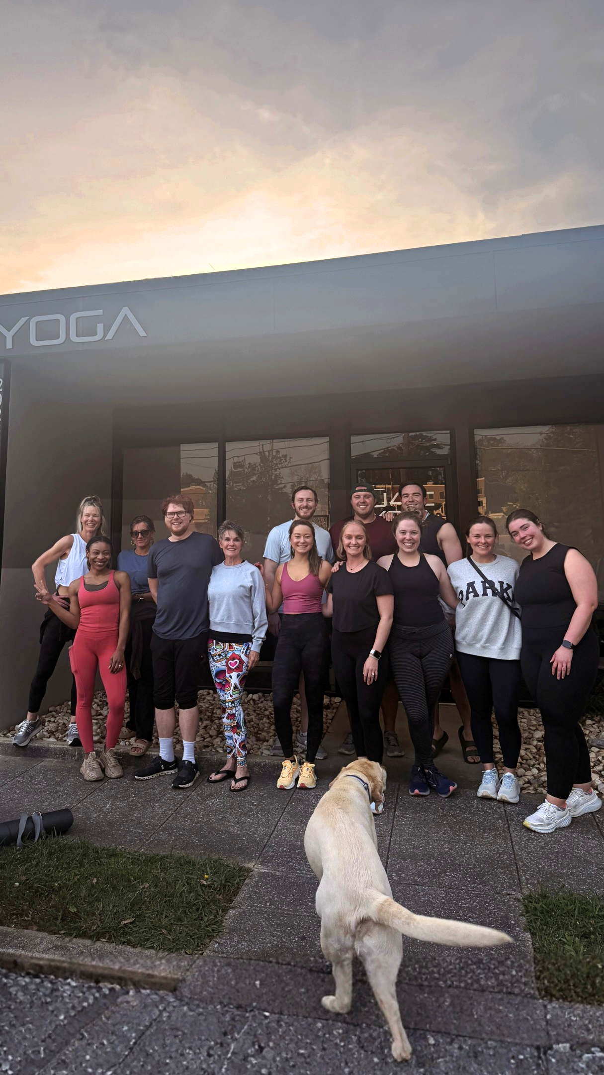 A group of fifteen people standing outside a building with glass doors, some smiling and posing for a photo. There is a yellow Labrador Retriever dog in the foreground, facing away from the camera. The building has a sign that appears to say 'YOGA' and is shown during the evening with a cloudy sky.