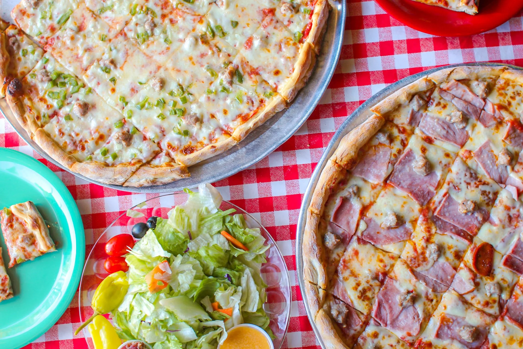 Two large pizzas on metal trays, one with green onions and cheese, the other with ham, sausage, and pepperoni, sitting on a red and white checkered tablecloth. In the foreground, there is a small plate of mixed salad with lettuce, cherry tomatoes, and yellow bell pepper slices, along with a small container of salad dressing.
