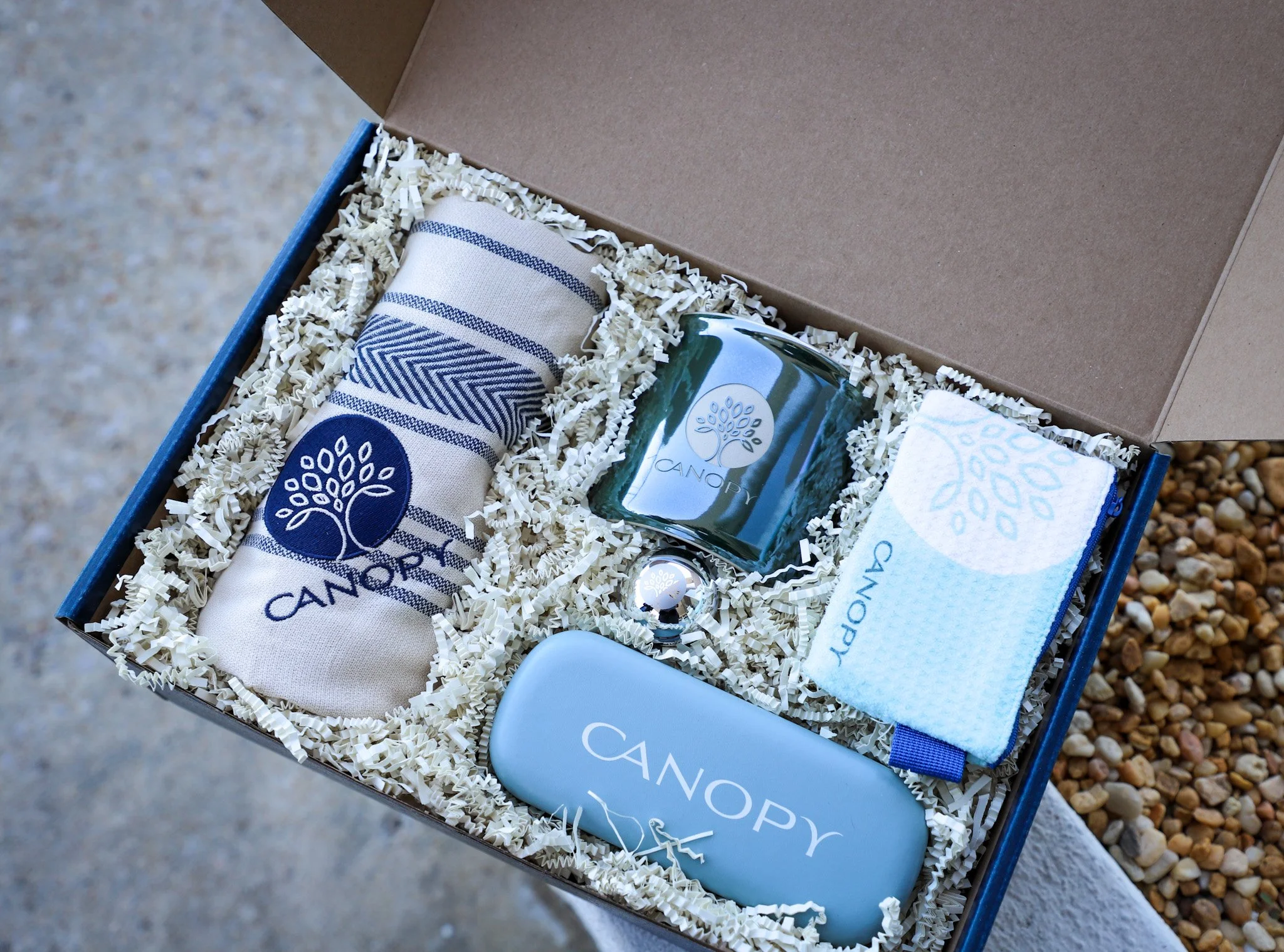 Open gift box with shredded paper filling, containing a folded striped cloth with a logo, a small green bottle with a logo, a white item with a logo, and a white cloth with a blue logo.