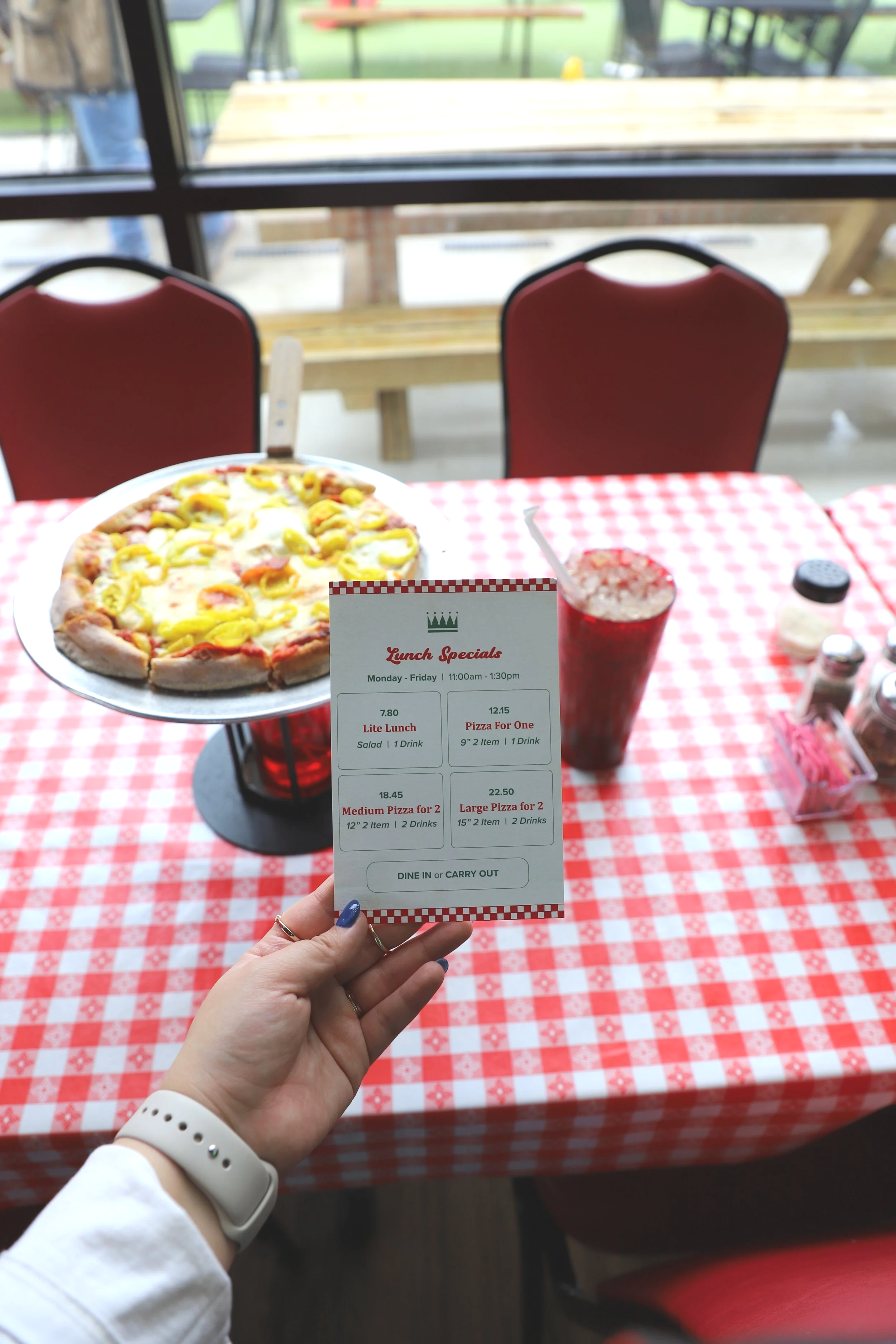 A person holding a pizza menu in front of a window at a restaurant table with a pepperoni and yellow pepper pizza on a metal tray, outside view of benches and a person walking.