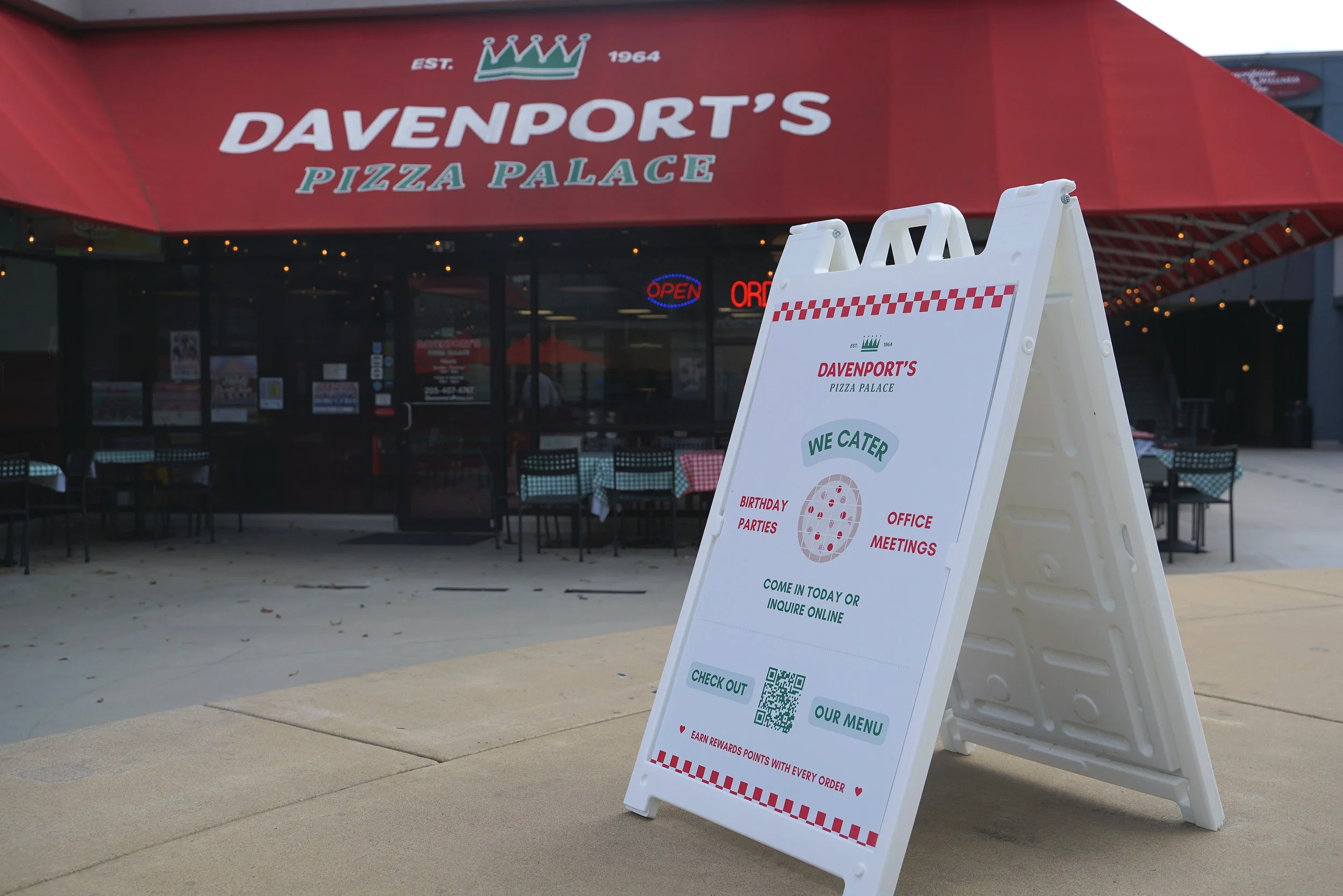 White A-frame sign outside Davenport's Pizza Palace restaurant with online ordering, rewards, and menu information, in front of a glass storefront with tables and chairs, red awning with the restaurant's name, and neon open signs.