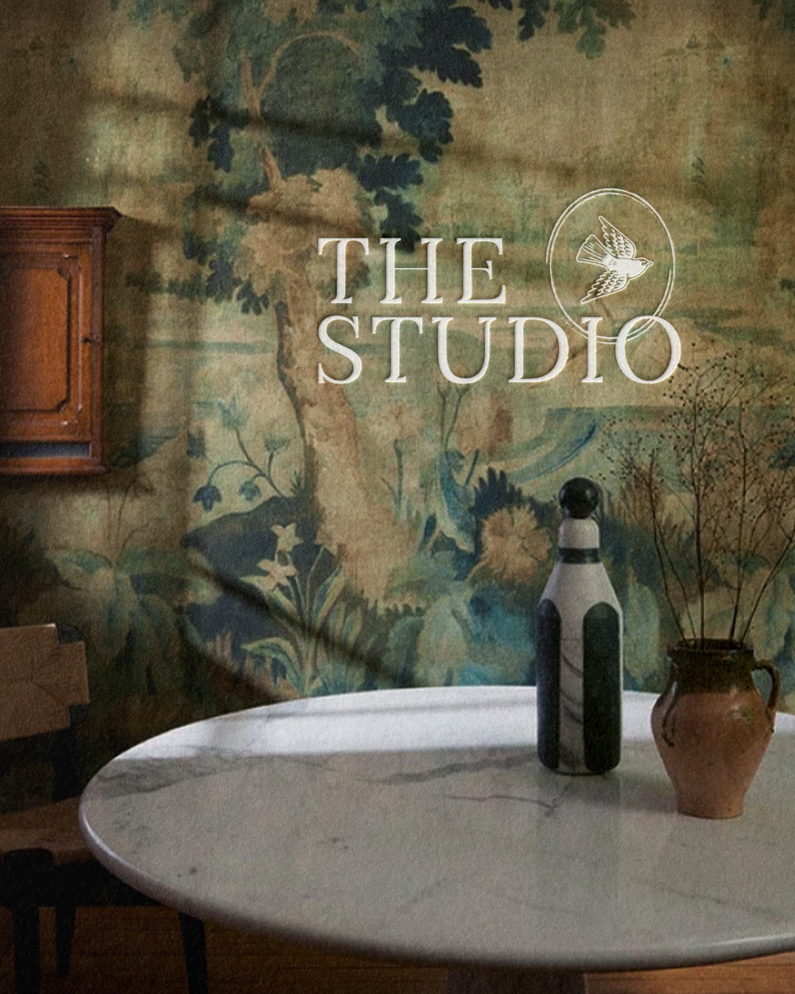 A round table with a dark top, decorated with a tall vase with painted black and white patterns, a smaller potted plant with dried branches, two bottles of oil or vinegar, and a mountainous painted wall in the background.