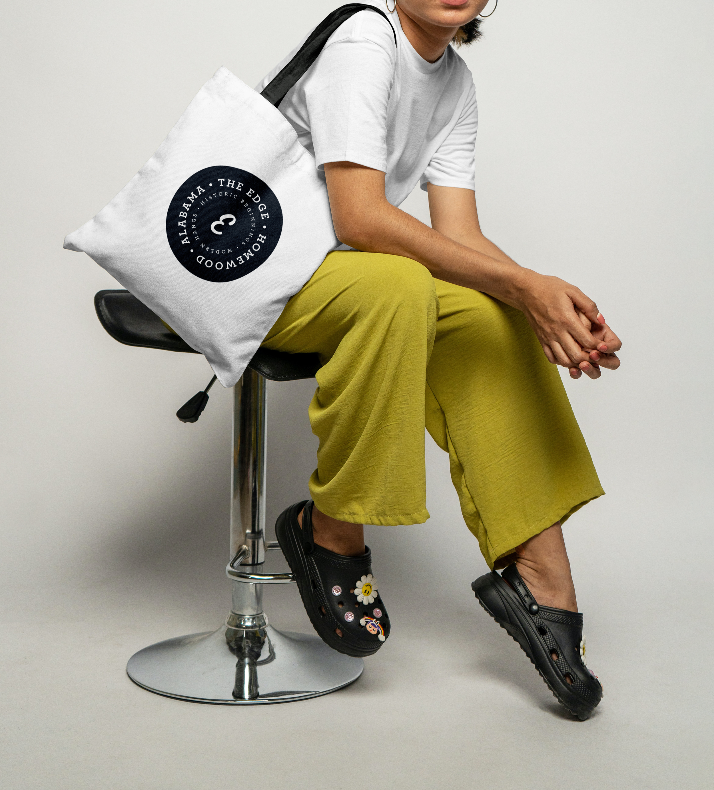 A person wearing a white t-shirt, yellow pants, and black sandals with floral and emoji decorations, is sitting on a black chair with a chrome base, with their hands clasped and legs crossed. The person has hoop earrings and is carrying a white tote bag with a black circular logo that reads 'ALLARAMA - THE EDGE, HOMEWOOD, MANH, HISTORIC BEGINNING, MODERN, HANGS'.