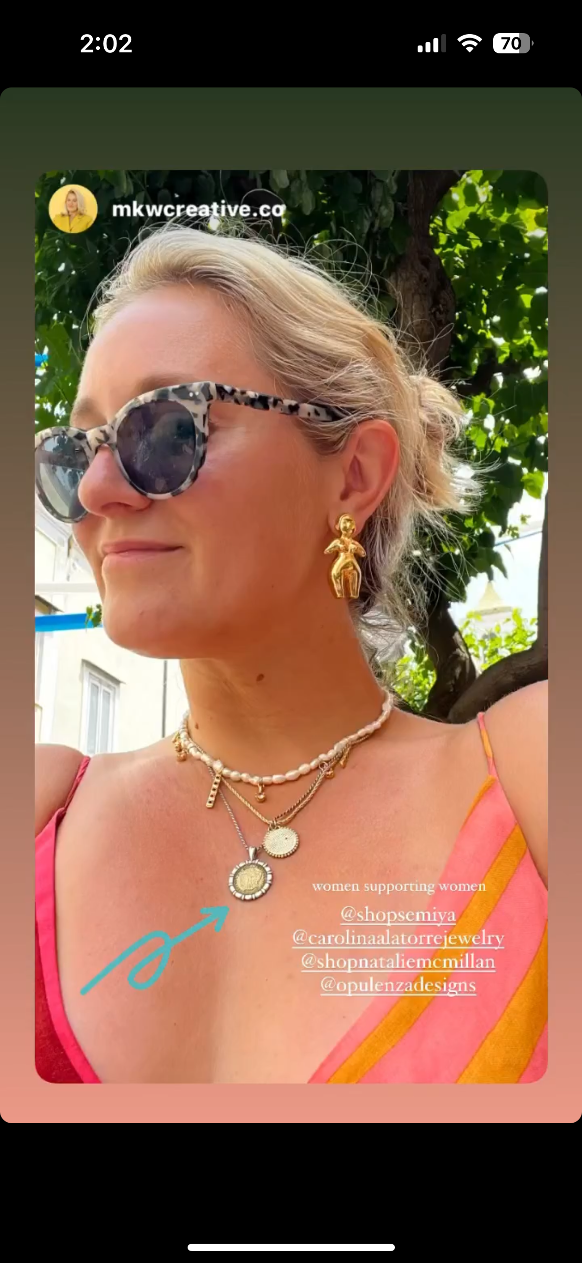 A woman with short blonde hair wearing patterned sunglasses, gold earrings, layered necklaces, and a pink and yellow striped top, smiling outdoors with trees in the background.