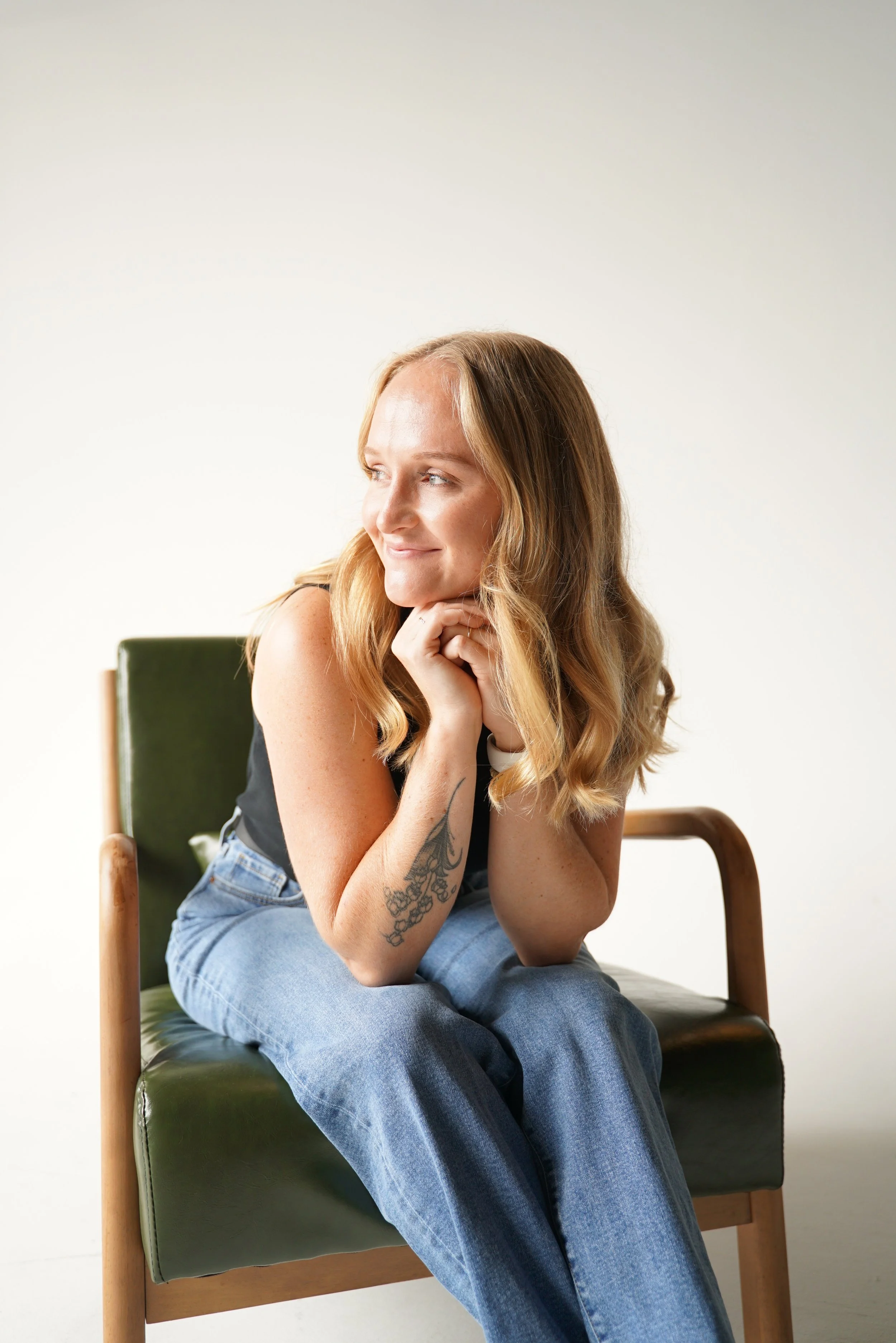 A young woman with long wavy blonde hair sitting on a green chair, resting her chin on her hand, smiling while looking to the side, against a plain white background.
