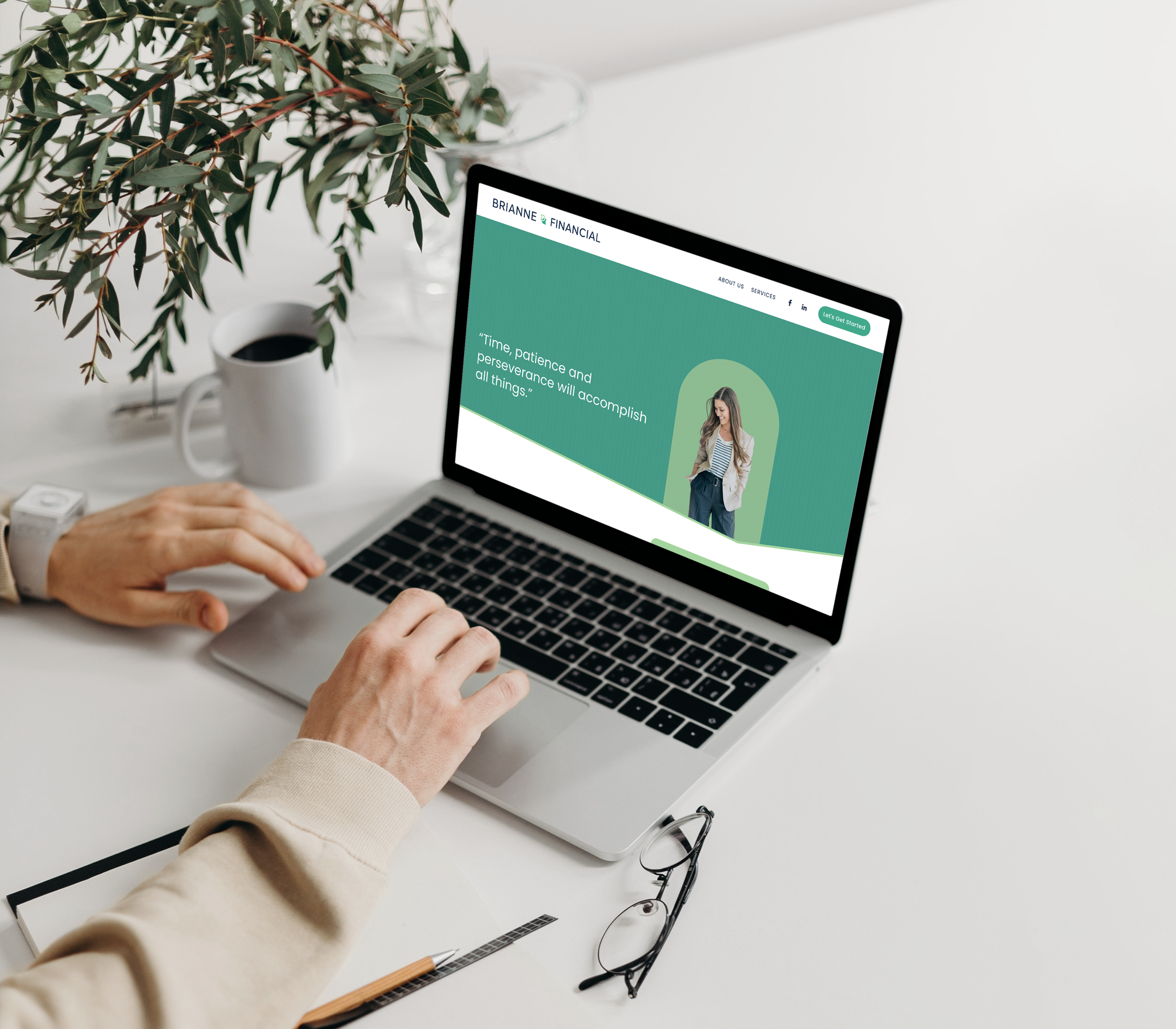 Laptop on a white desk showing a website for Brianné Financial with a woman in a blazer, greenery in the background, next to a cup of coffee, eyeglasses, a notebook, and a pen