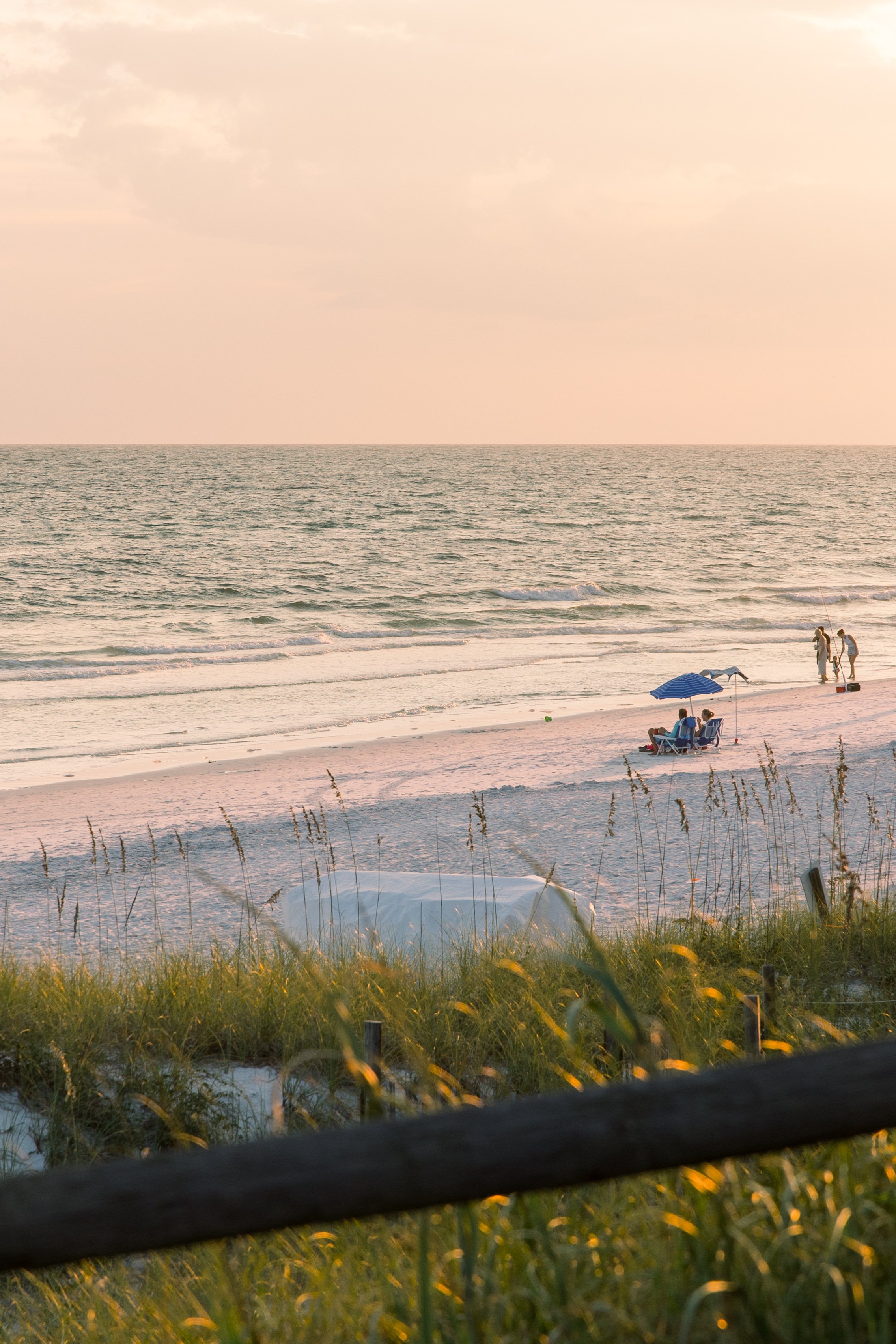 View of the ocean with a sandy beach, a blue umbrella, and people walking along the shore during sunset. Foreground has grass and a wooden fence.