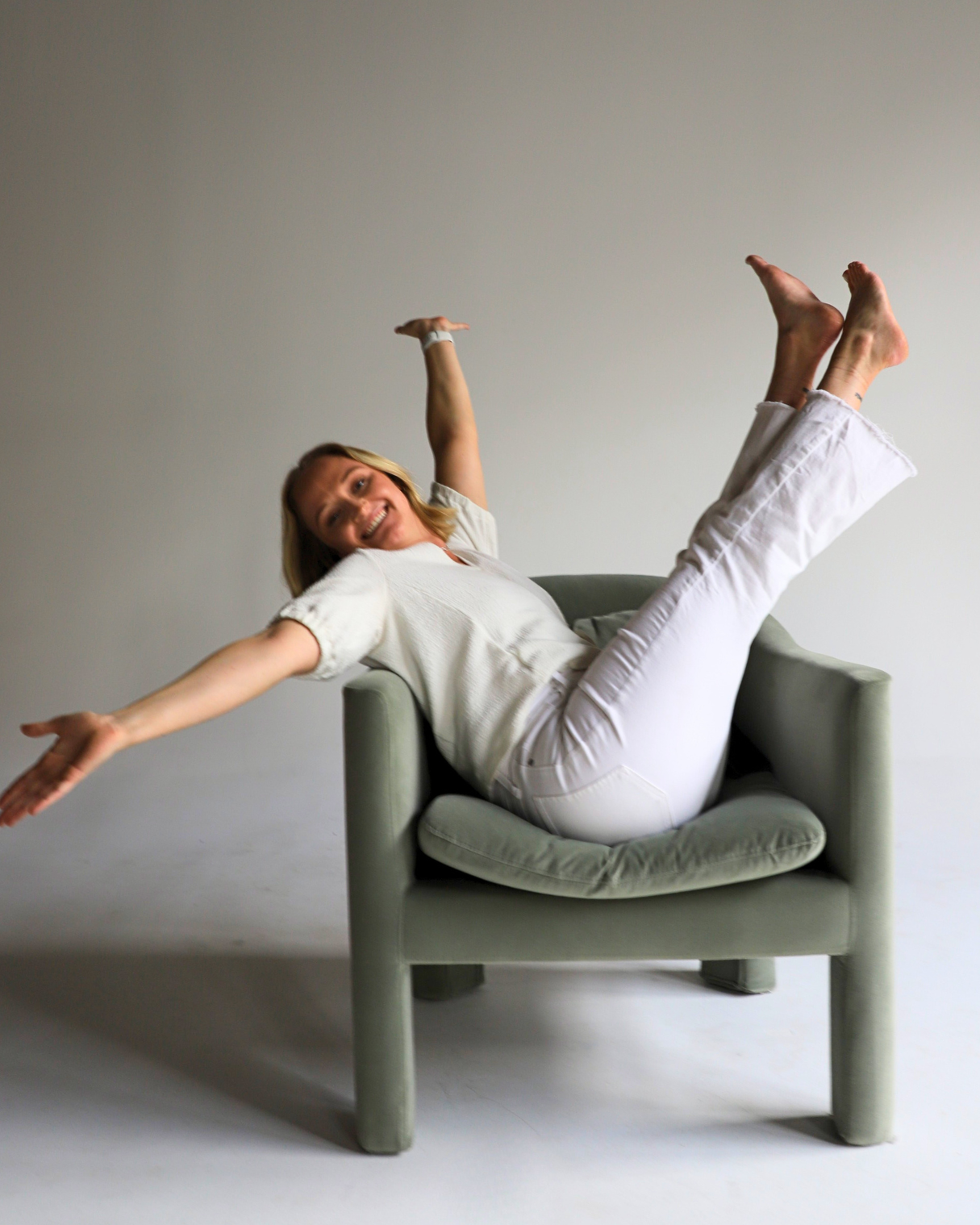 A woman in white pants and a beige shirt sitting in a gray armchair, smiling, with her arms and legs extended outward, appearing to fall or lean back in a joyful pose.
