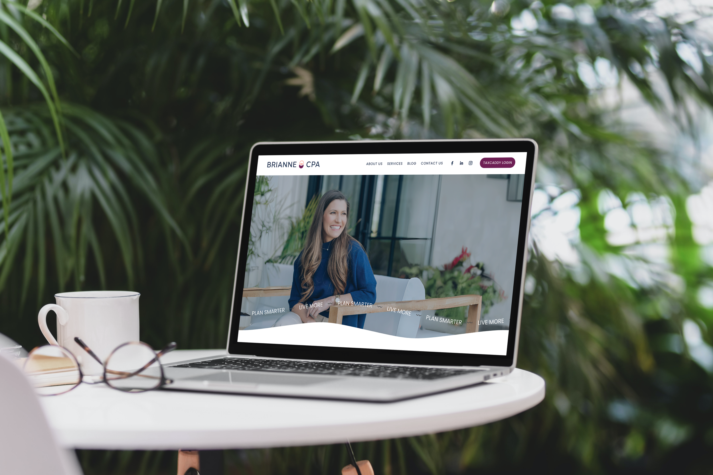 A laptop on a white table displaying a woman's smiling face on a website, surrounded by green foliage. A white mug, an open notebook, and eyeglasses are also on the table.