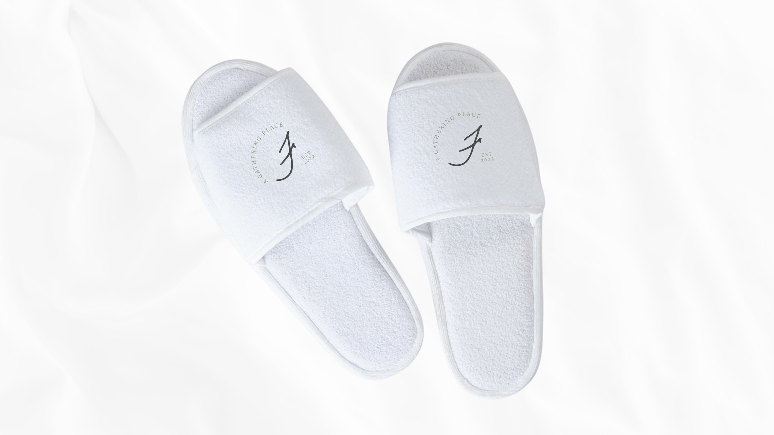 White slippers with folded straps, featuring a black logo and the text 'A Gathering Place' along with 'Est 2022' on the straps.