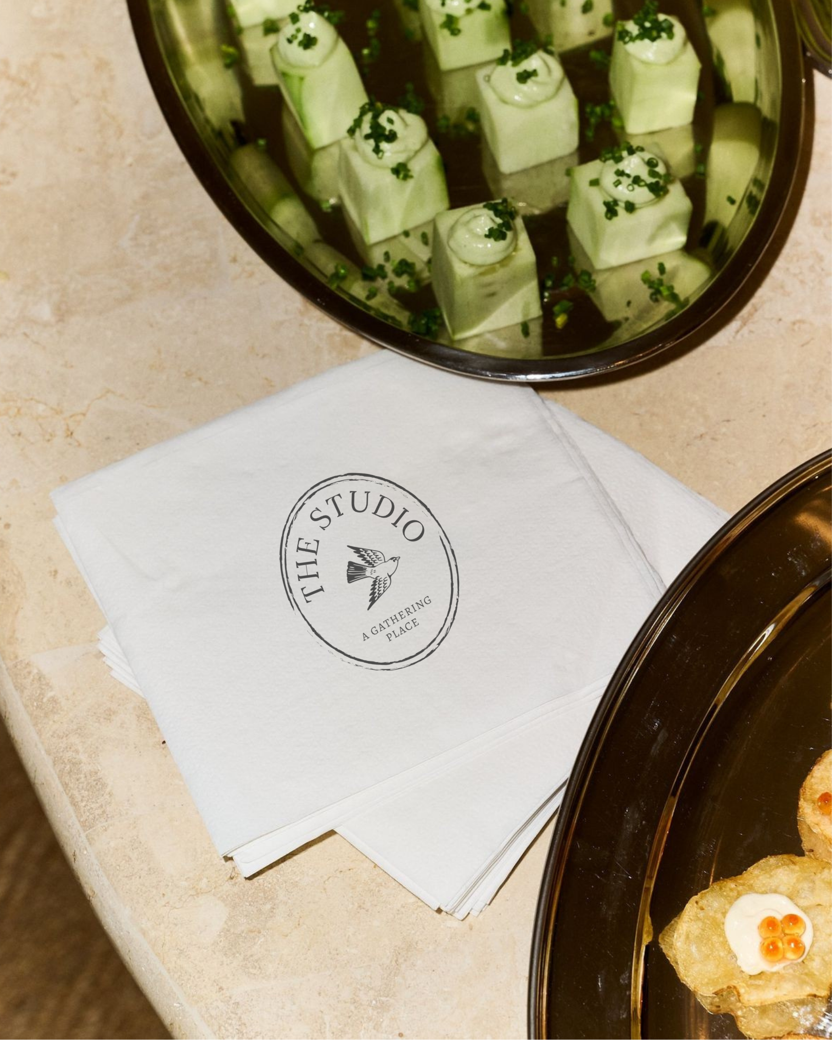 Close-up of a plate of deviled eggs garnished with chopped chives and black pepper, next to a white napkin with a logo and a black tray with more food in the background.