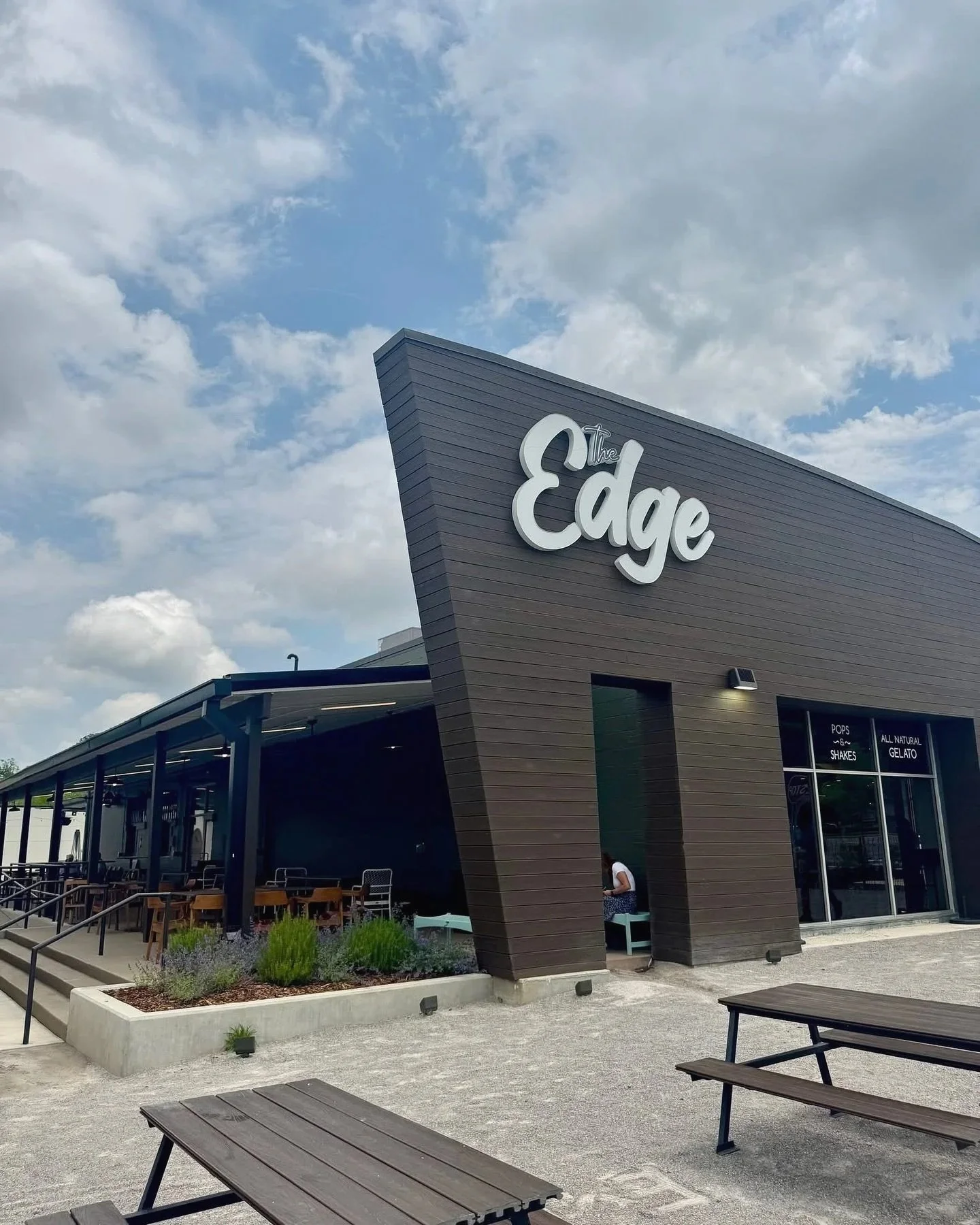 Exterior of The Edge building with a modern dark brown facade and a prominent white sign. Outdoor seating area with tables and benches, landscaped with plants, under a partly cloudy sky.