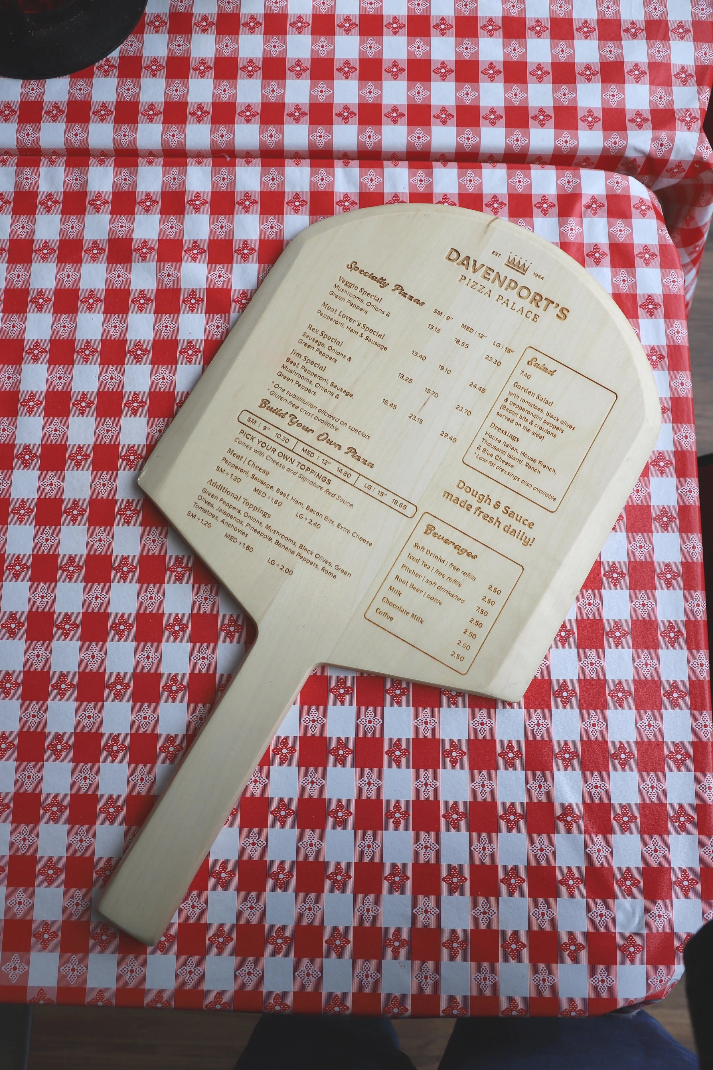 A bakery or pizzeria menu on a wooden paddle resting on a red and white checkered tablecloth with a floral pattern. The menu features various special pies, salads, dressings, and beverages.
