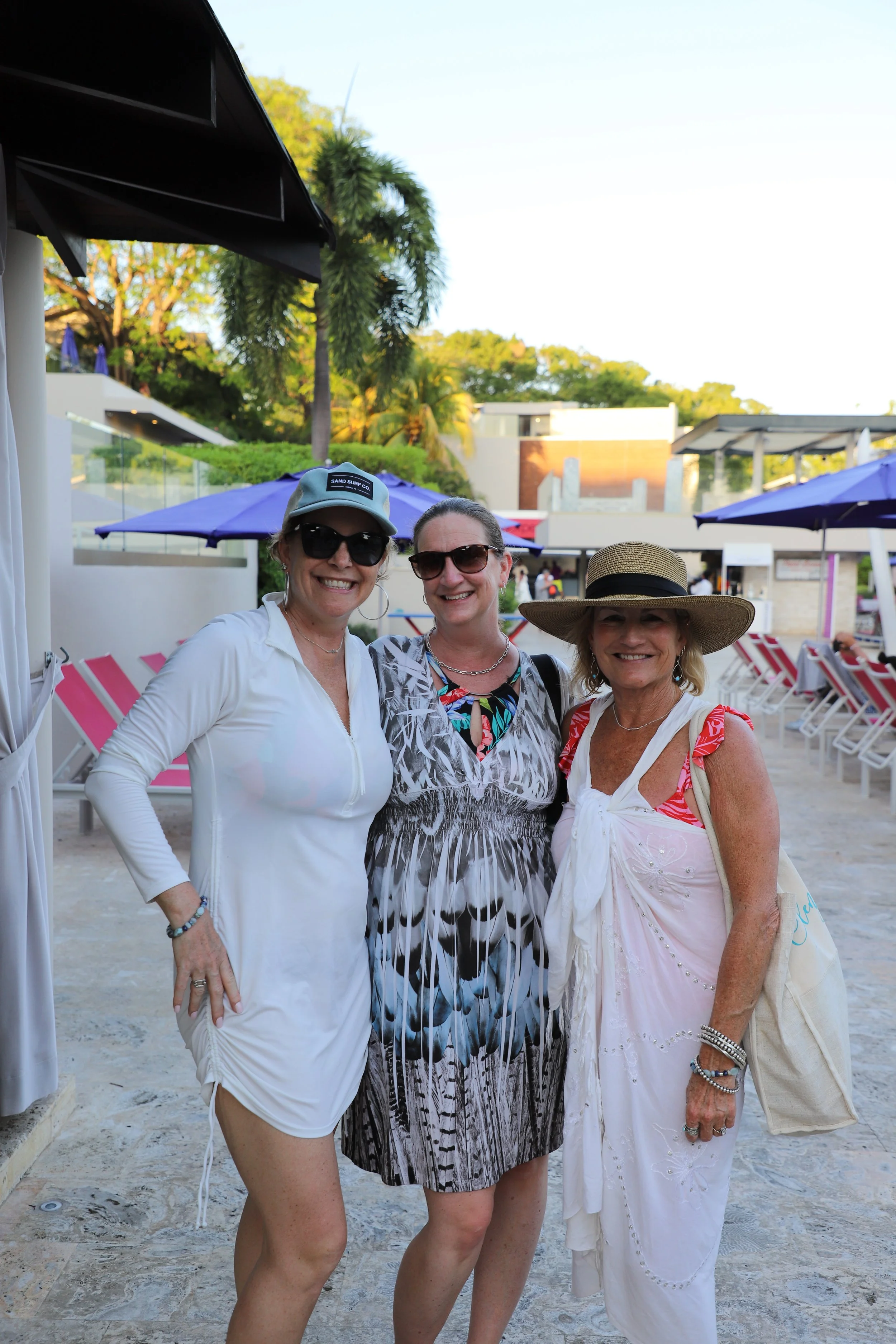 Three women smiling at an outdoor gathering, wearing summer clothing and hats, standing on a sandy area with umbrellas and chairs in the background, with trees and modern buildings behind.