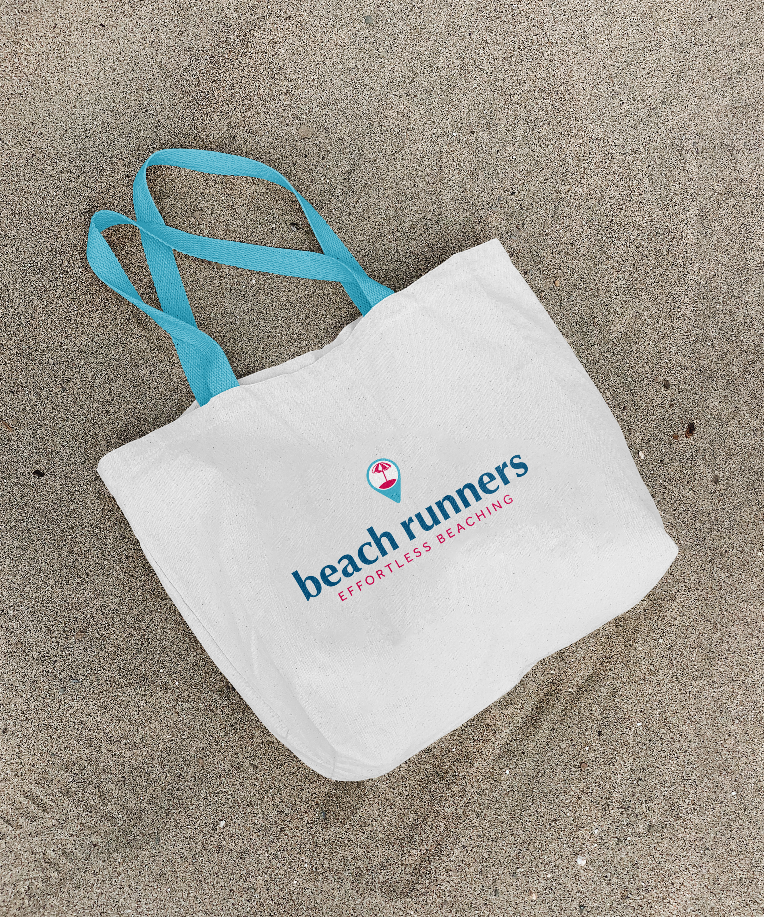A white shopping tote bag with blue straps lying on sandy beach. The bag has the words 'beach runners' printed on it with a pink umbrella icon.