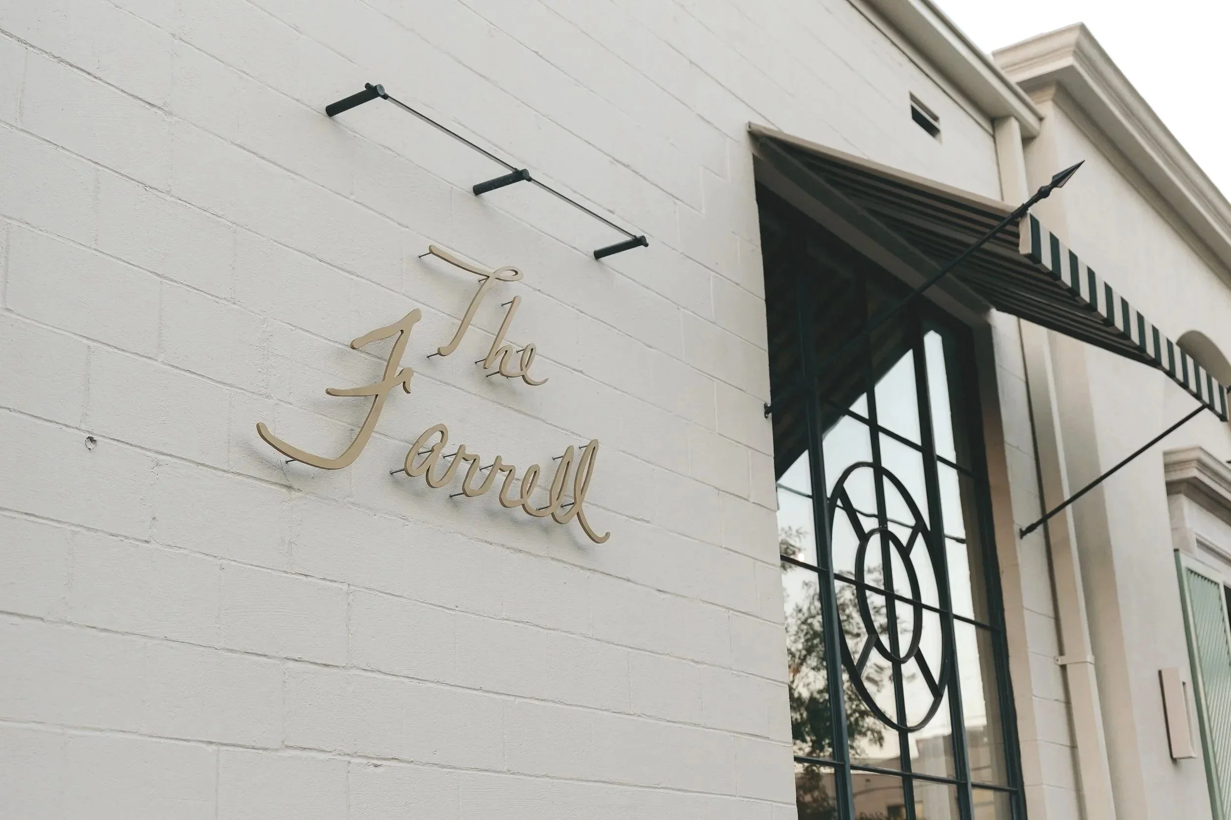 Close-up of a white brick building exterior with elegant cursive gold sign that reads 'The Jarcelle', with a black and white striped awning and decorative black metal window grille.