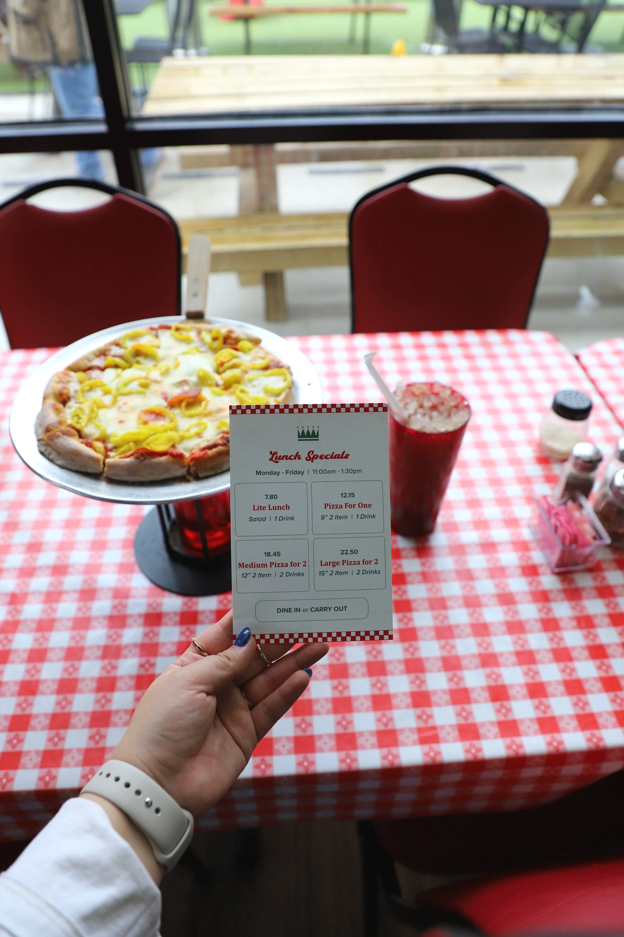 A person holding a restaurant menu with lunch specials listed, placed on a red and white checkered tablecloth. The background shows a pizza on a tray, a drink, and a window with chairs and tables outside.