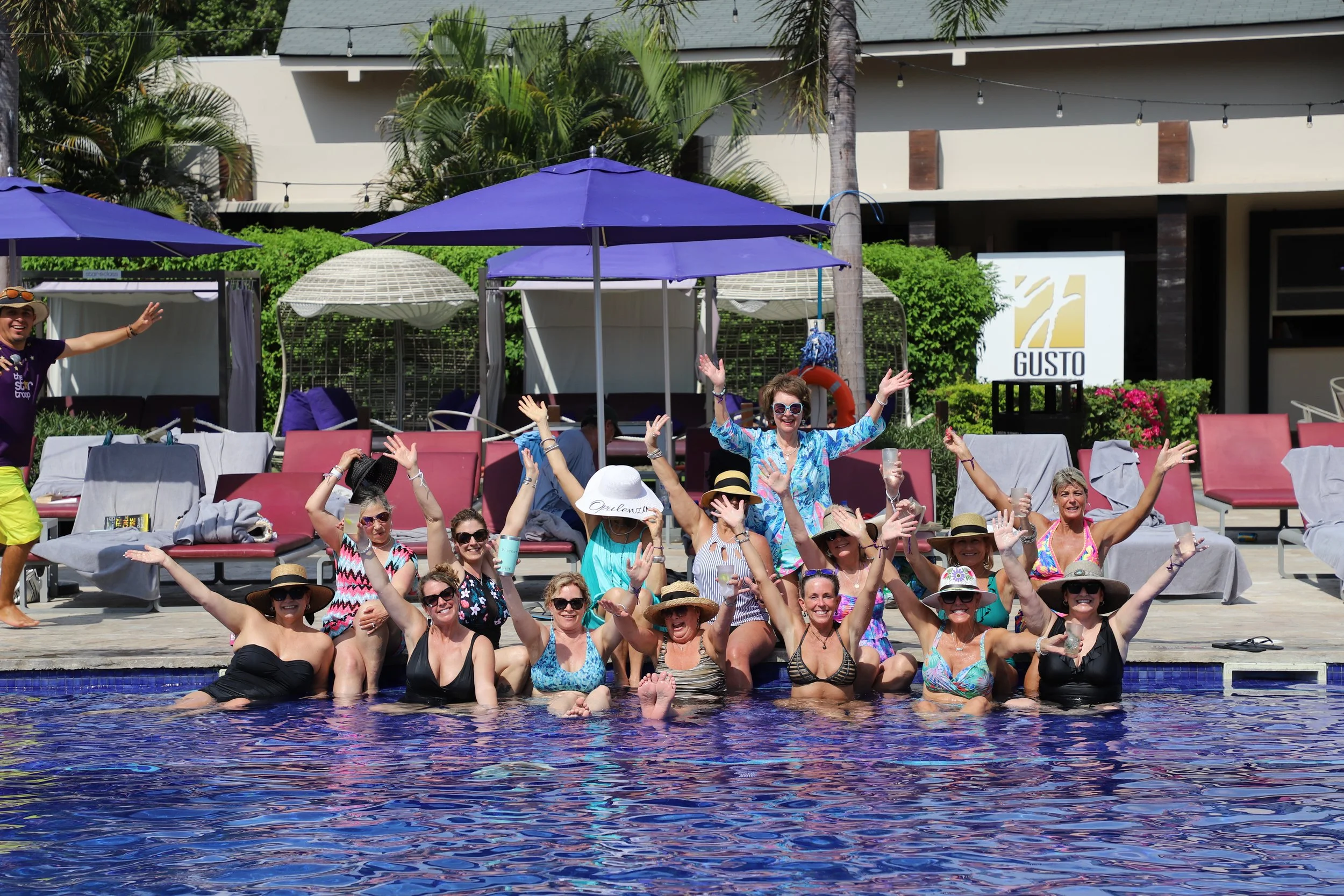 Group of people enjoying a pool party in swimwear, with some holding drinks and raising their hands, surrounded by poolside lounge chairs, umbrellas, and tropical greenery.