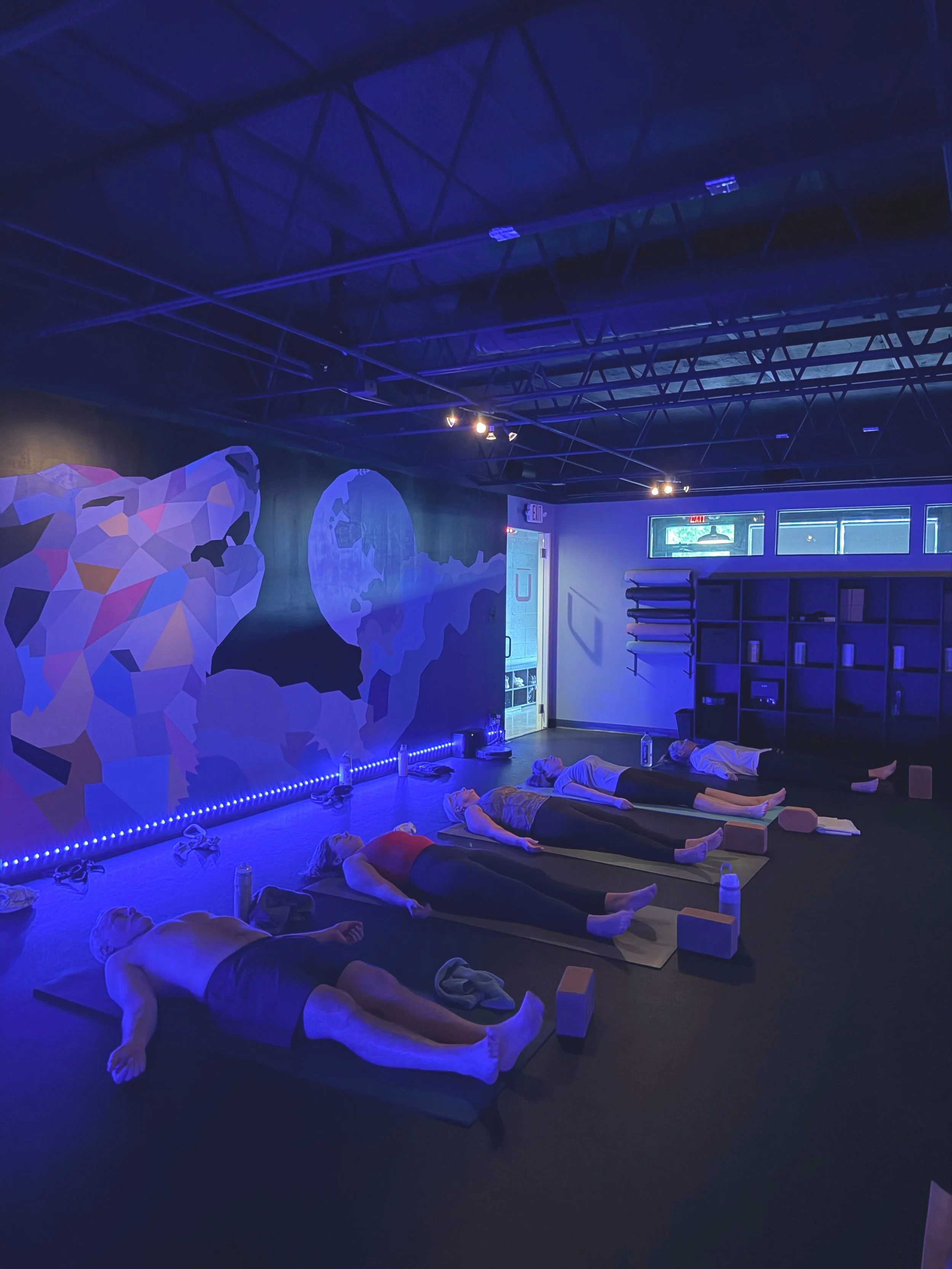 A group of people lying on yoga mats in a dark room with blue lighting, likely participating in a relaxation or meditation session. There are wall murals of a polar bear and the earth, an exit door, and exercise equipment in the room.