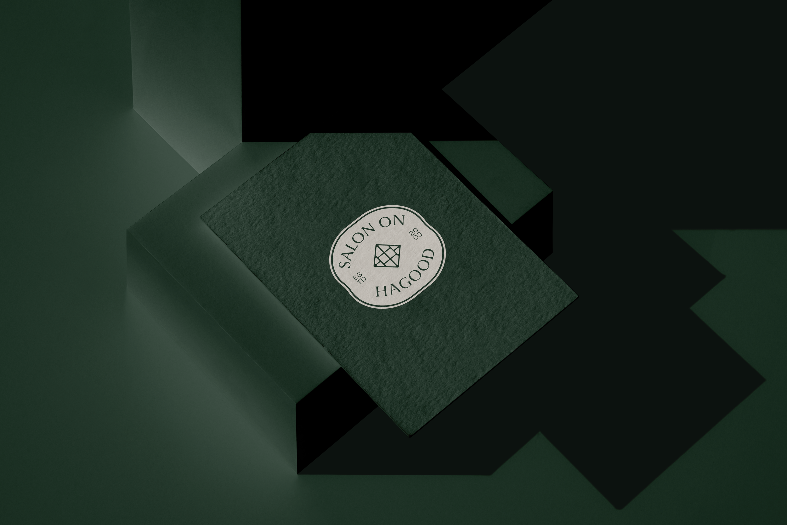 Dark green textured box with a white circular label reading 'Salón Hogood' and decorative border, placed on a black surface with green shadows.