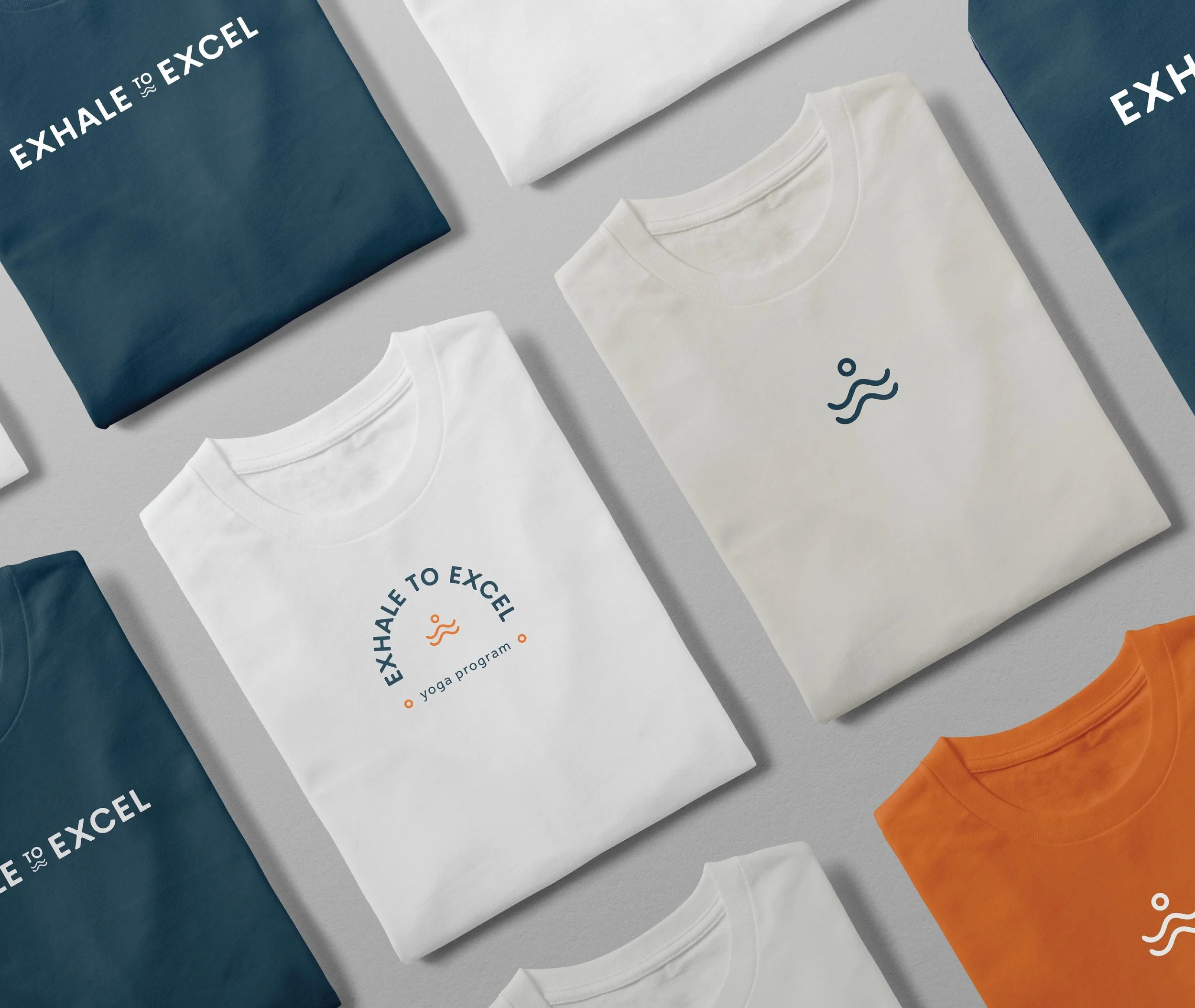 White T-shirts with a yoga logo, blue and white boxes labeled "EXHALE TO EXCEL", orange pants, and white clothing tags displayed on a light gray surface.