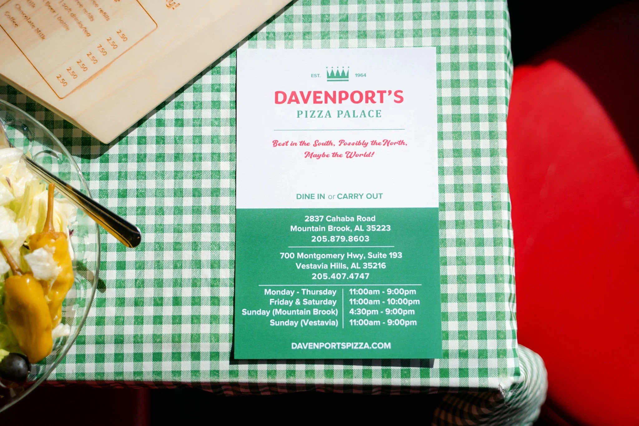 Close-up of a menu or flyer for Davenport's Pizza Palace with address and hours of operation, placed on a green and white checkered tablecloth, with part of a salad bowl visible on the left.