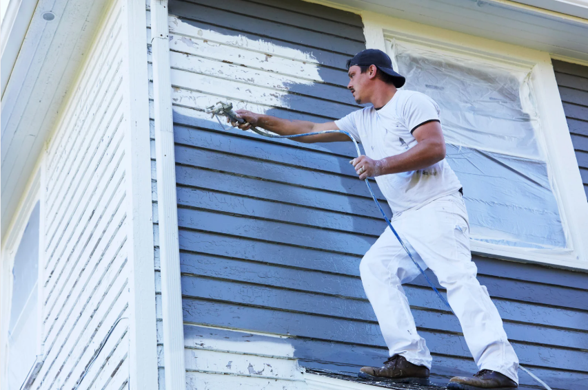 Man painting the exterior of a house blue, standing on a scaffold, wearing white clothes and a black cap.