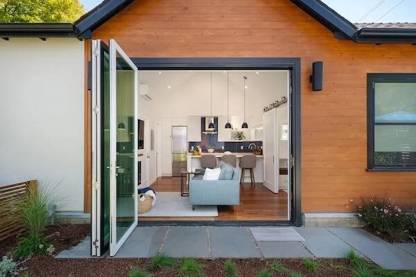 Open sliding glass door leading into a modern living room with a gray sofa, kitchen with white cabinets, black backsplash, and pendant lights, and a dining area with four beige chairs.