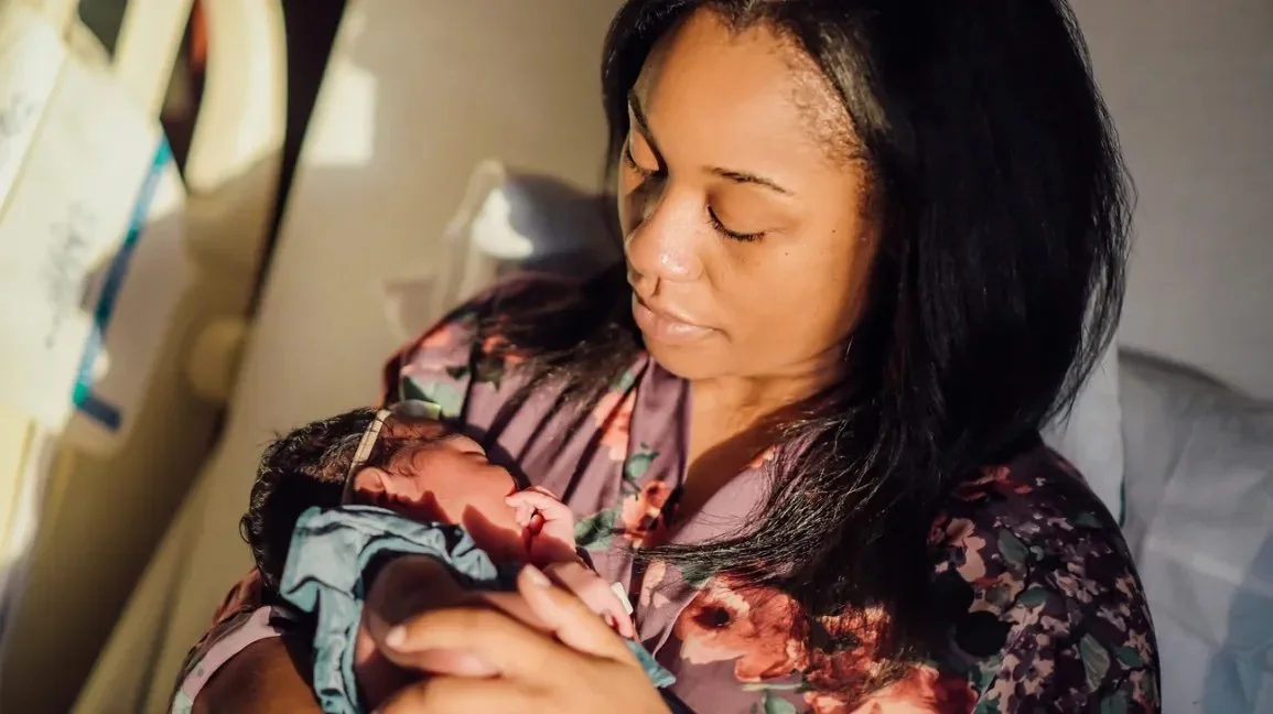 A Black woman holds a newborn baby in a hospital room, looking down at the baby tenderly.