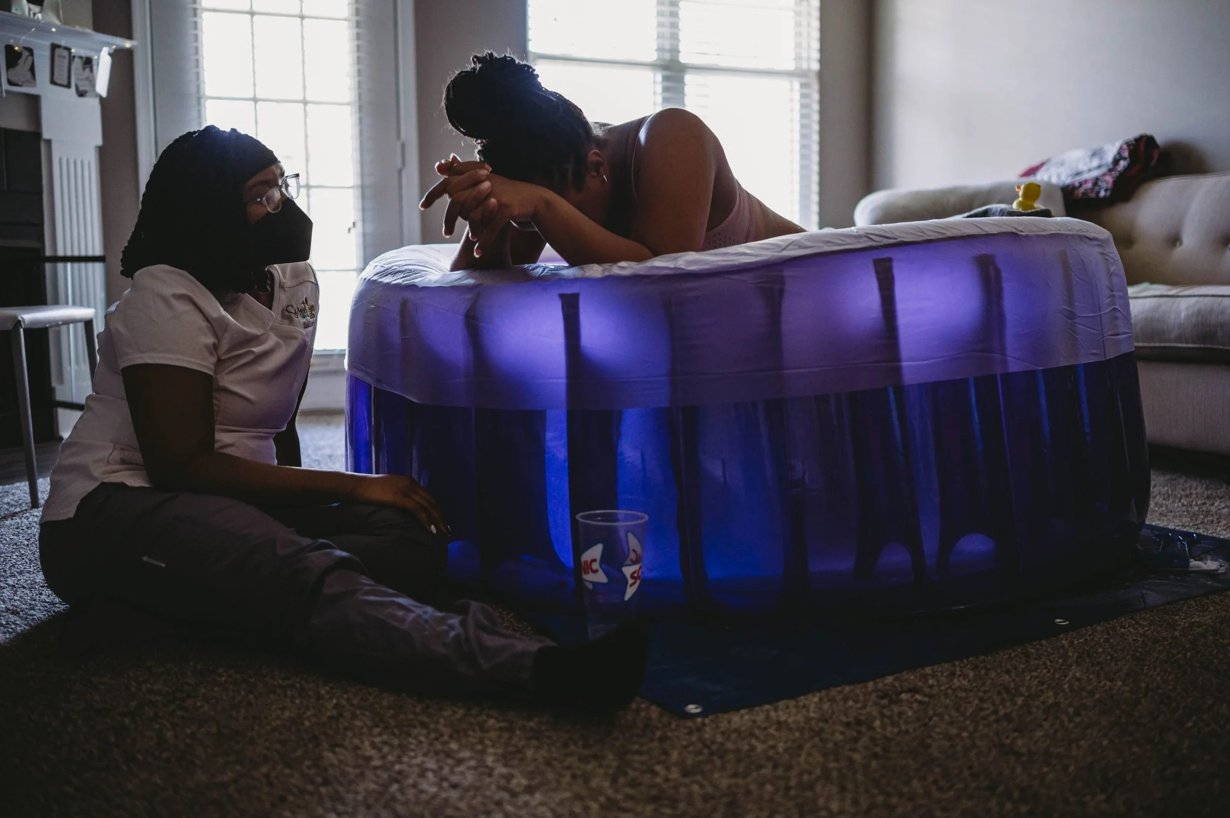 A woman labors in a birthing tub with support from a doula wearing a face mask.