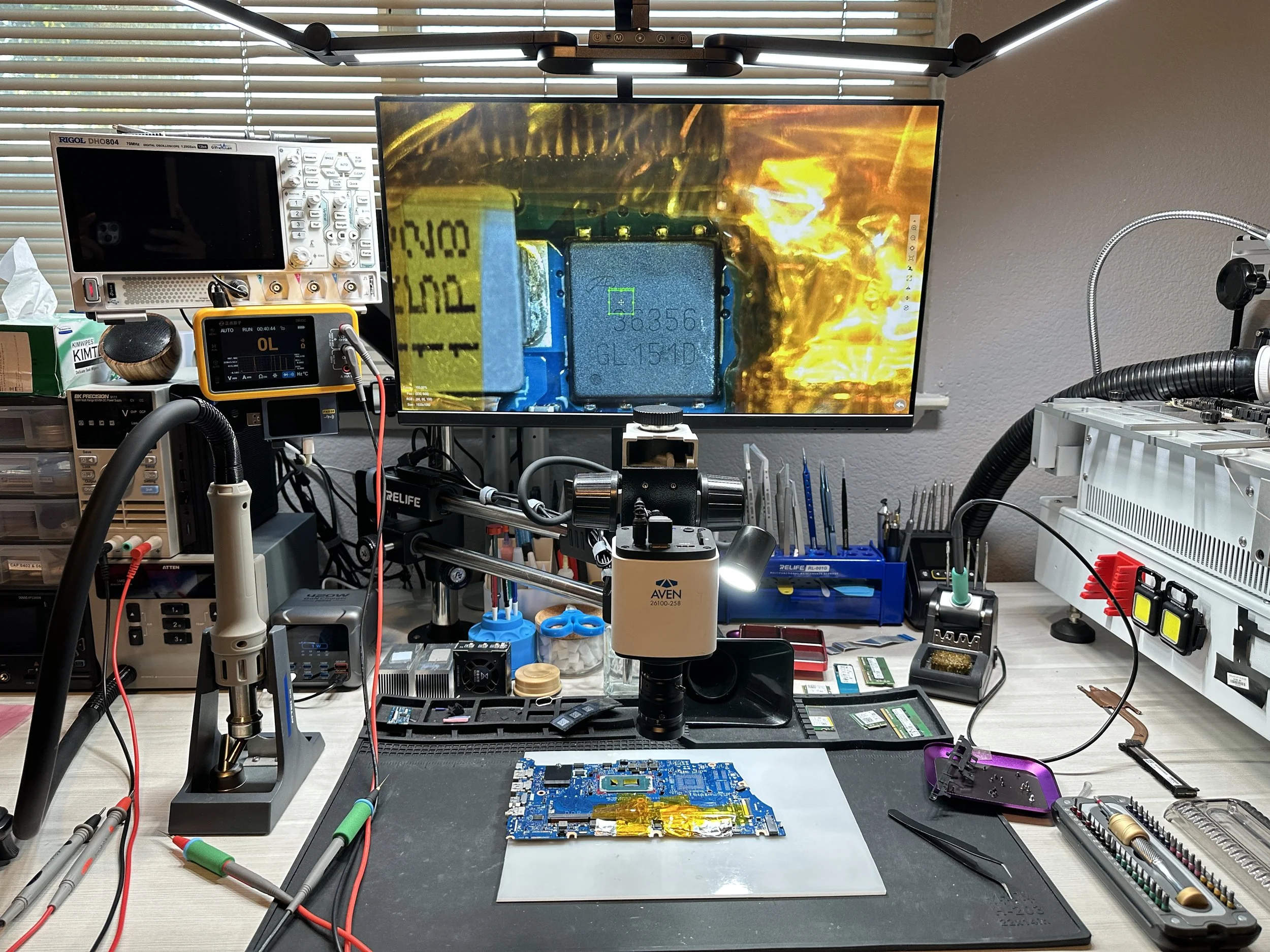 Electronics repair workspace with a circuit board, tools, and testing equipment on a cluttered workbench.