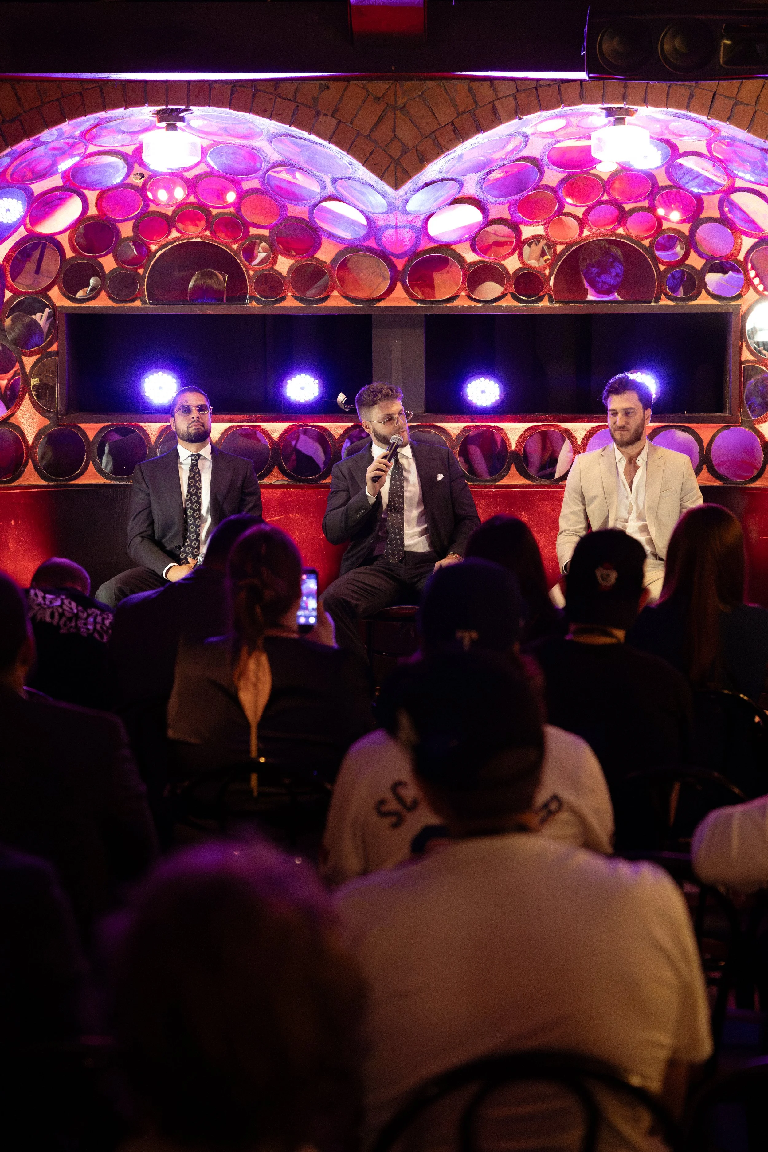 Three men seated on a stage, with one man in the middle holding a microphone, in front of an audience. The background features a decorative wall with circular mirrors and colorful lights.