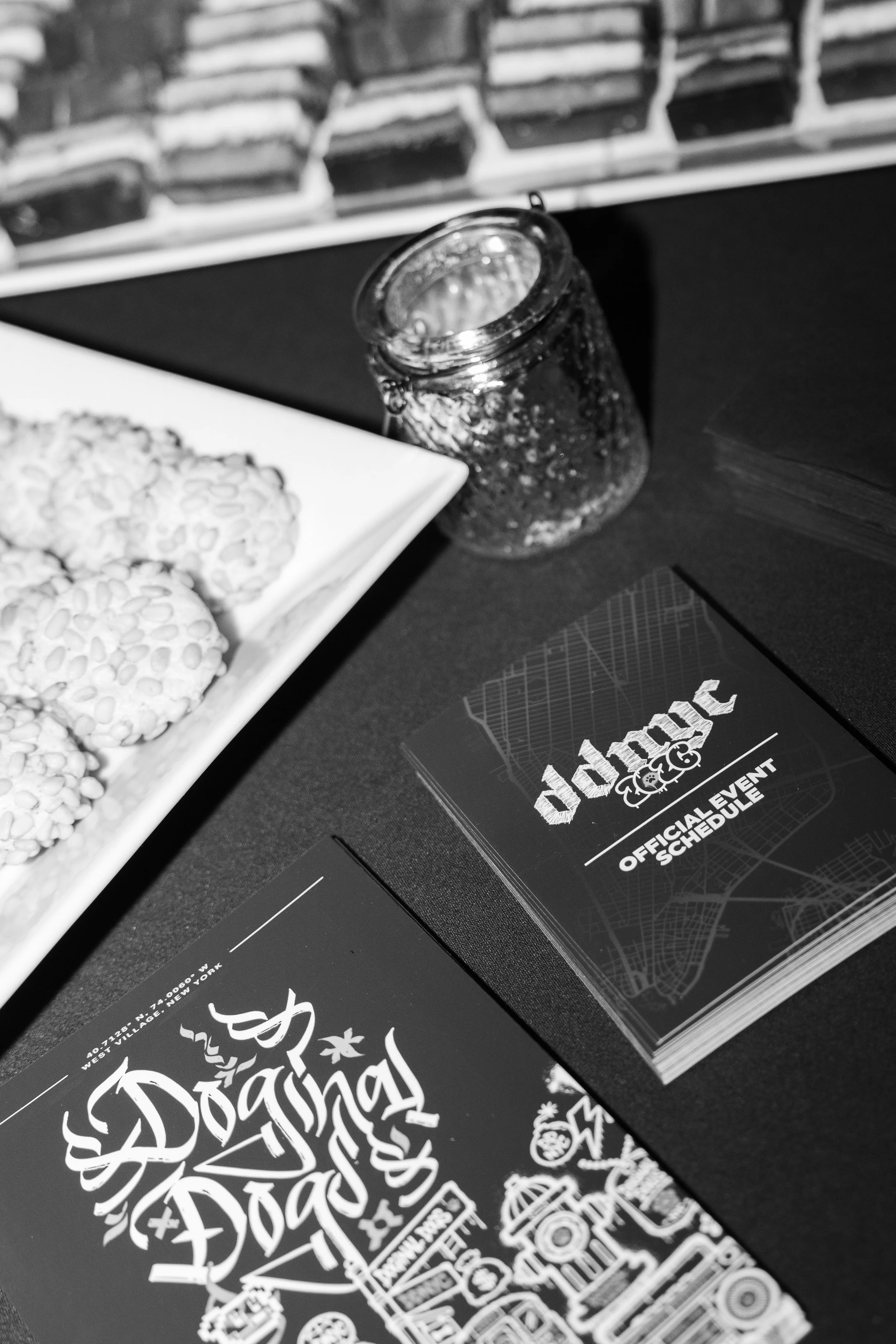 Black and white photo of merchandise with graphic design and text, including a menu or schedule titled 'Official Event Schedule', and a deck of playing cards or illustrated cards on a dark surface.