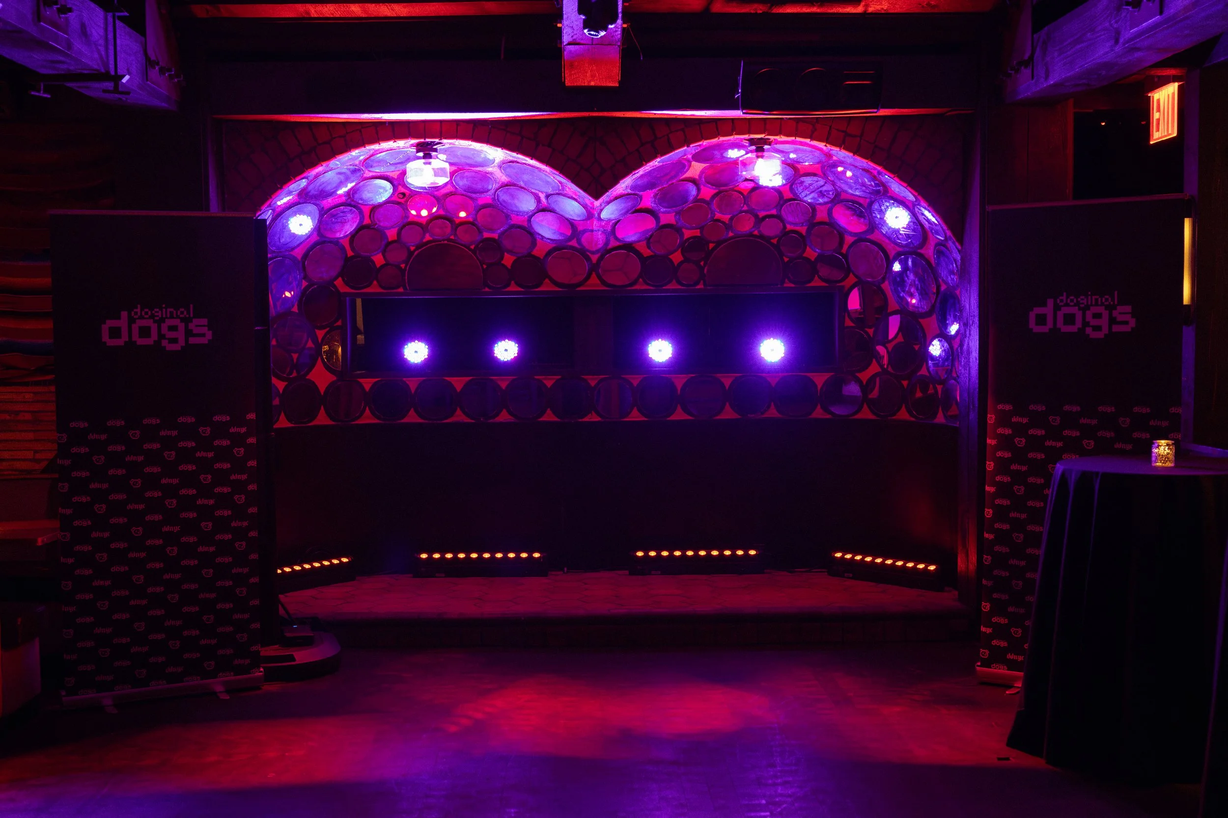 A dark indoor space with a DJ booth illuminated by purple and pink lights, featuring the logo 'doginal dogs' on two black banners on each side of the booth, with a brick wall backdrop and dancing floor area in front.