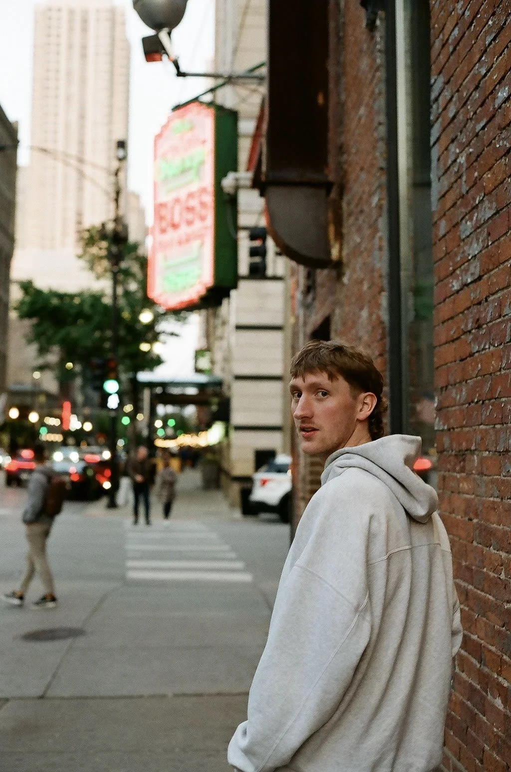 A young man in a gray hoodie standing against a brick wall on a city street at dusk, with blurred pedestrians and cars in the background and a neon sign hanging above.