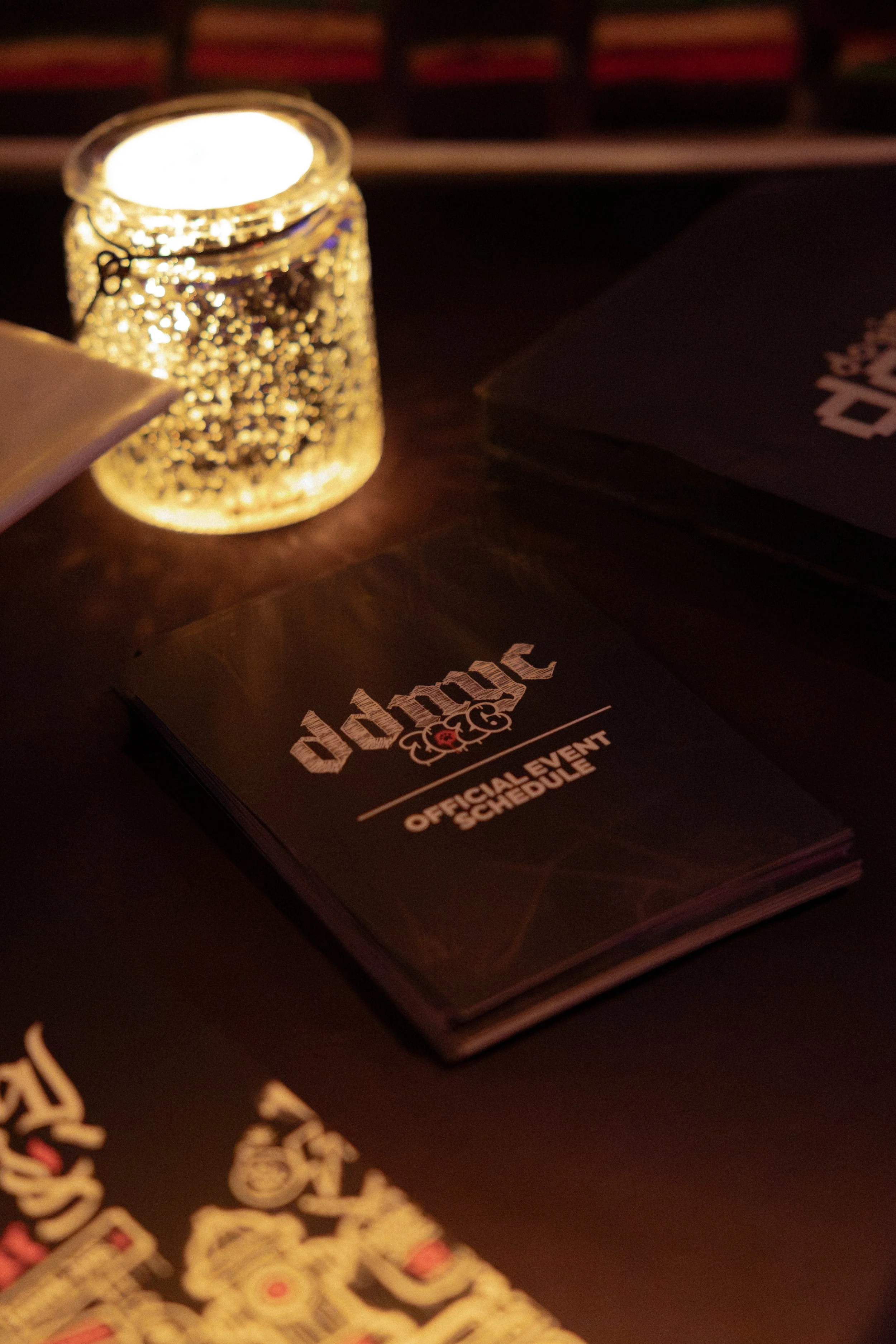 A glowing candle inside a decorative glass holder with gold accents, placed on a dark surface next to a black booklet or menu with the word 'douche' printed on it.