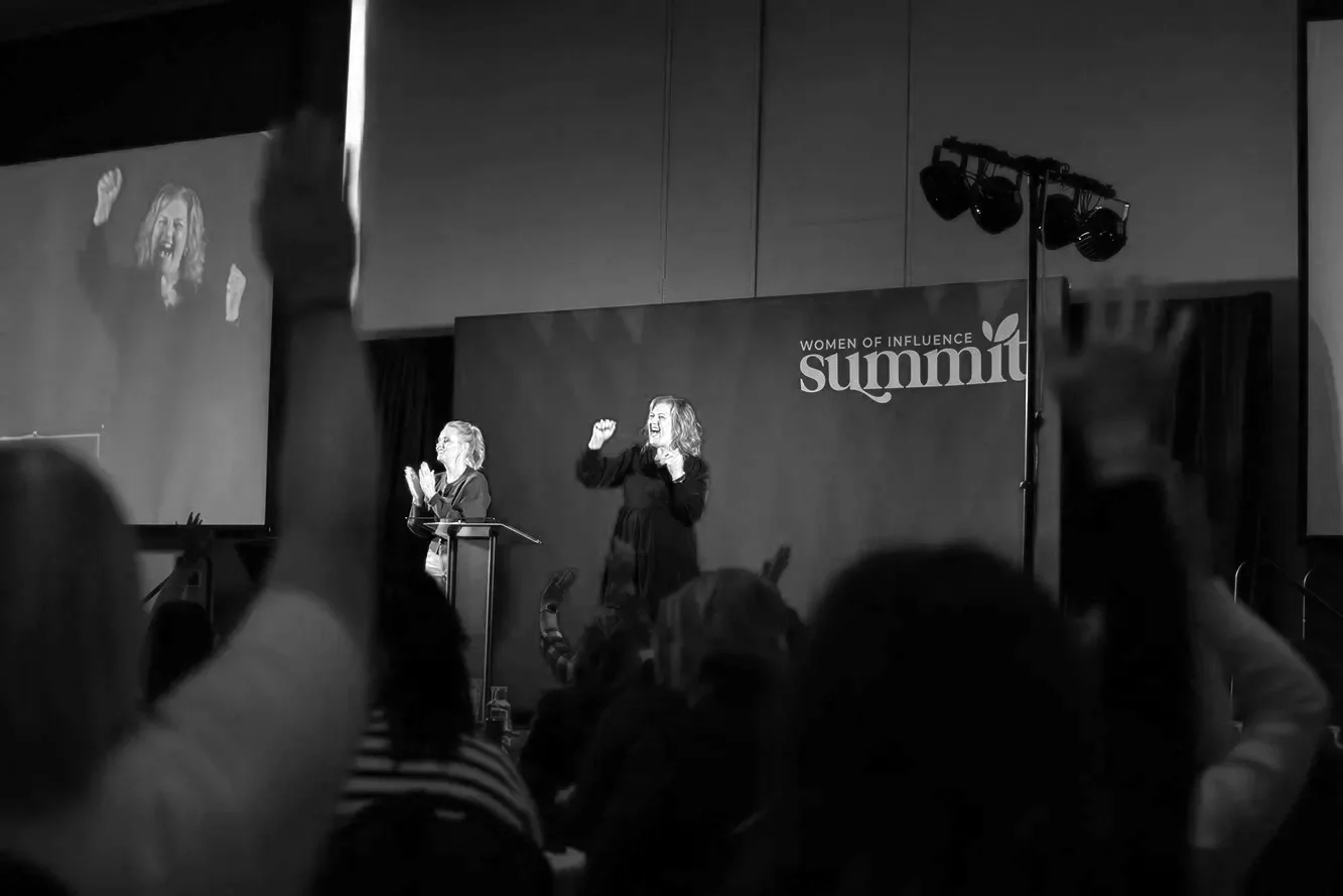 Black and white photo of a woman speaking or presenting on stage at the Women of Influence Summit, with a large screen displaying her image and audience members raising their hands.