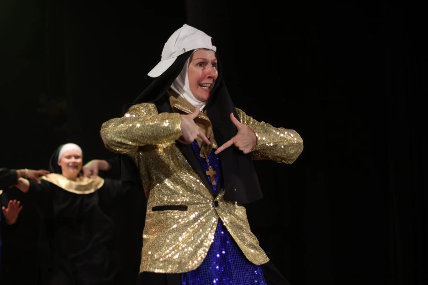 A woman dressed as a nun in a gold sequin jacket, black habit and White backwards cap, performing on stage with others in similar costumes, gesturing confidently with her hands and smiling.