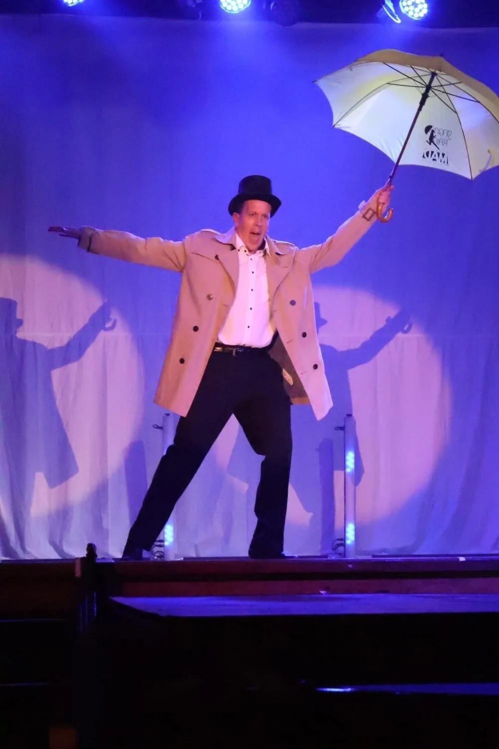 Performer on stage wearing beige trench coat, black top hat, and holding an umbrella, with arms outstretched, under stage lights.