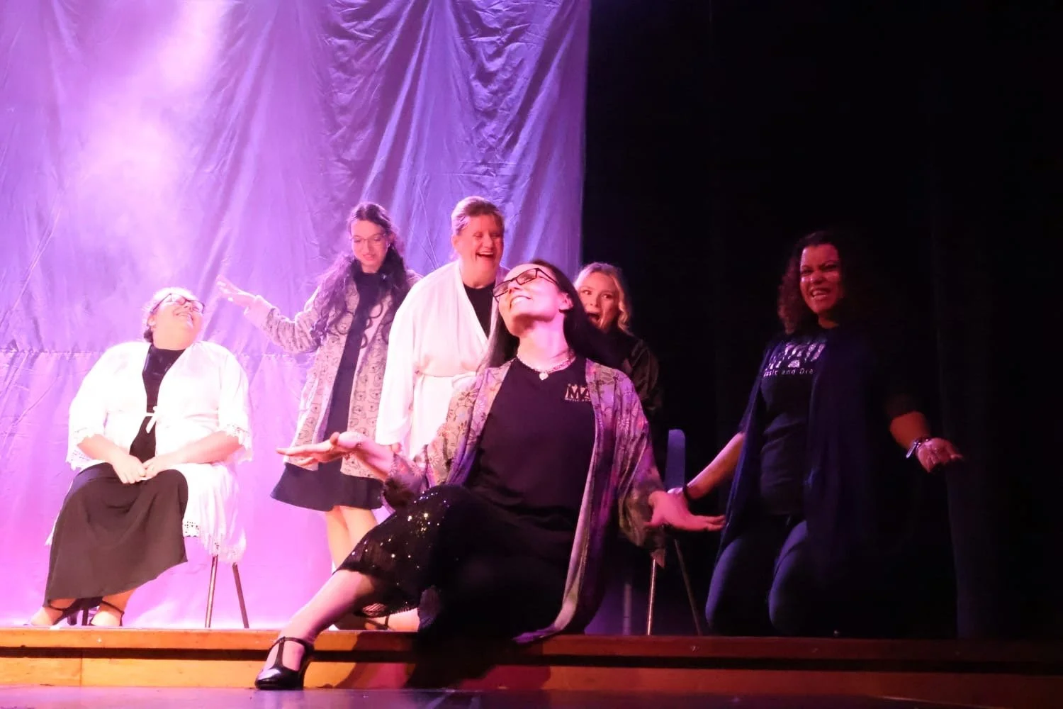 A theatrical cast on stage, with six women posing and smiling, one seated and five standing, in front of purple stage curtains, under stage lights.