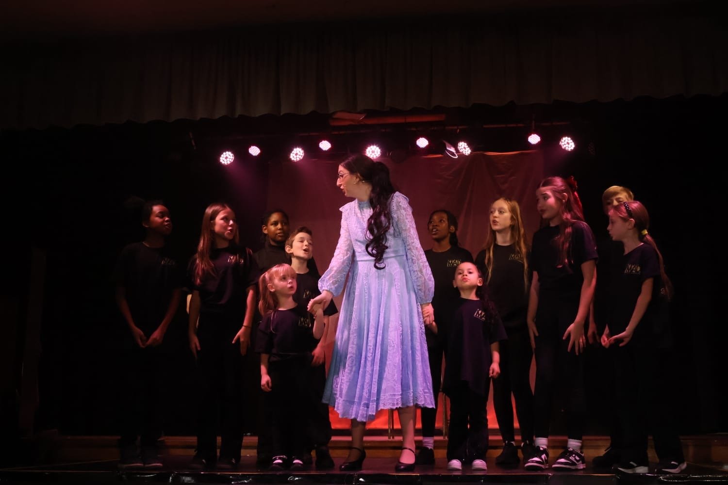 A group of children on stage with a woman in a long dress, holding hands, during a performance or show, with stage lights overhead.