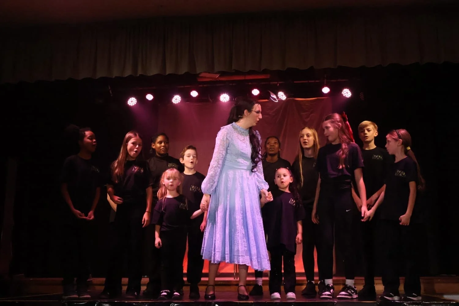 A woman in a light blue dress holding hands with young girls on stage with singing group in black shirts.