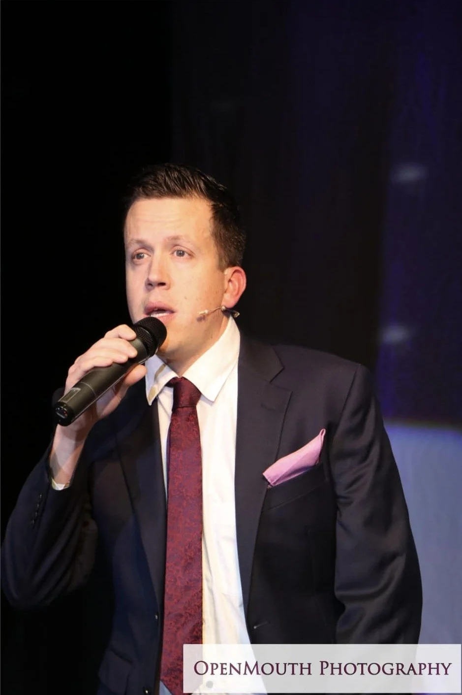 A man in a dark suit, white shirt, and red tie holding a microphone and speaking on stage, with a dim background and stage lighting.