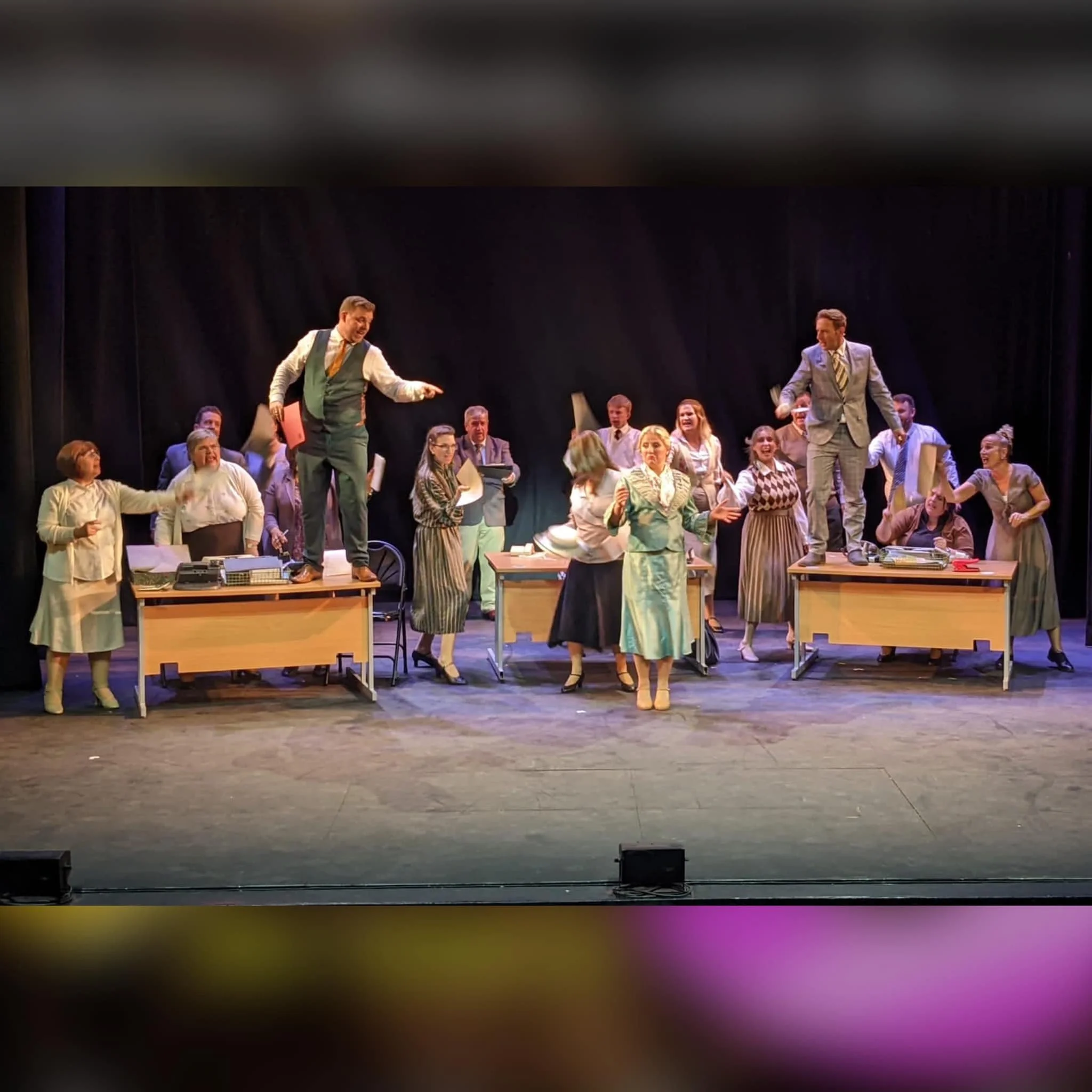 The image shows a theater scene with actors performing on stage, dressed in vintage office attire, with some standing on desks and engaging in a lively performance.