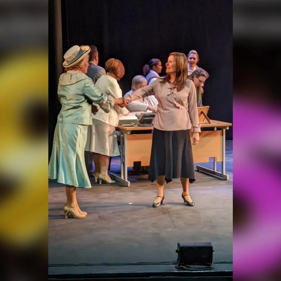 Woman in beige blouse and dark skirt shaking hands with an older woman dressed in a light-colored suit and hat on stage during a theatrical performance, with several other actors and a desk in the background.