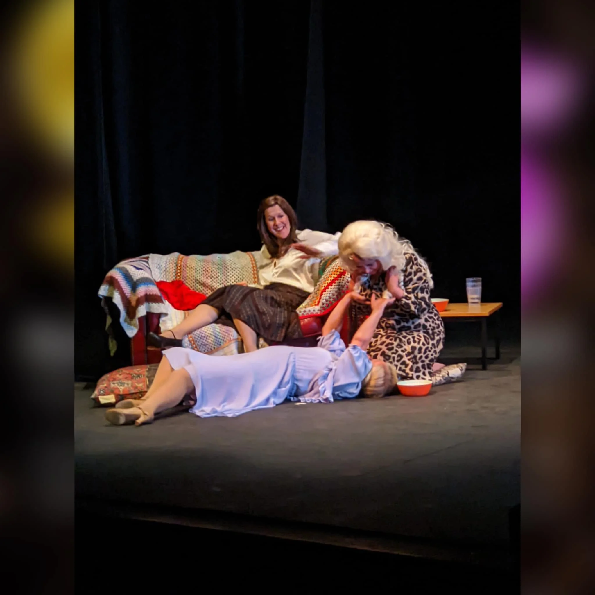 Three women in vintage clothing engaging in a playful scene on a stage, with one lying on the floor, one sitting on a couch, and the other kneeling, all laughing and interacting.