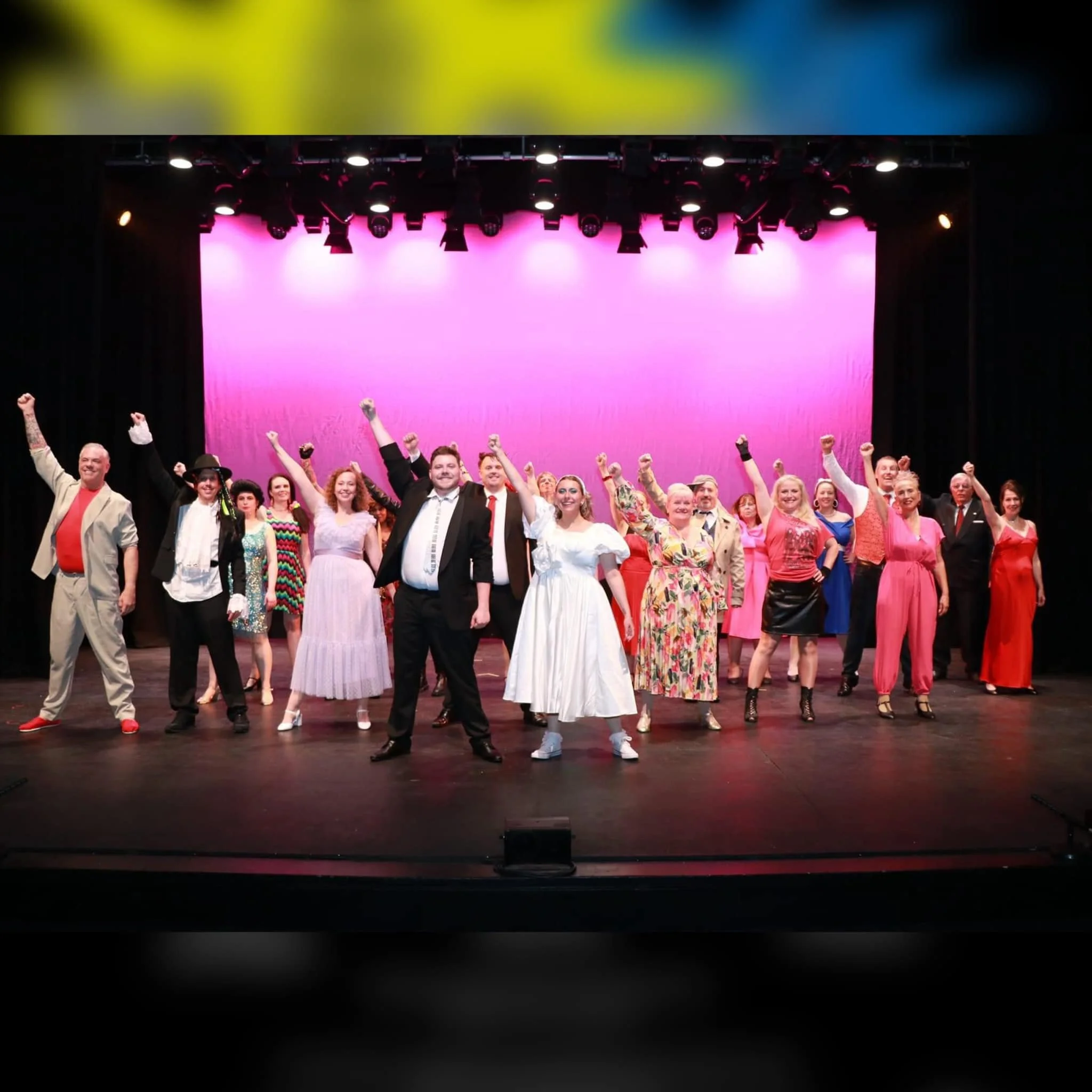 A group of diverse people on stage, posing and raising fists in a performance or celebration, with pink lighting and a pink backdrop.
